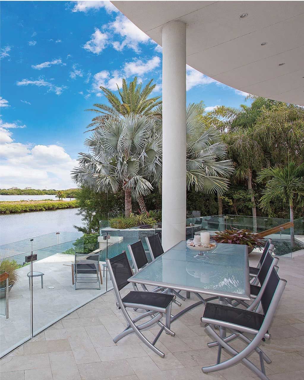 Outdoor patio with glass table and chairs overlooking a river. Palm trees and blue sky.