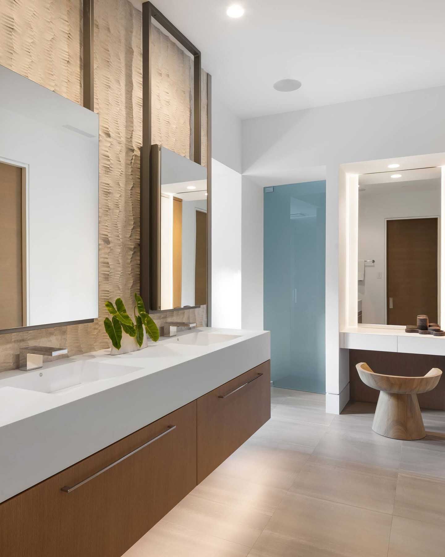 Modern bathroom with double sinks, mirrors, and wood cabinetry. Neutral tones with textured wall.