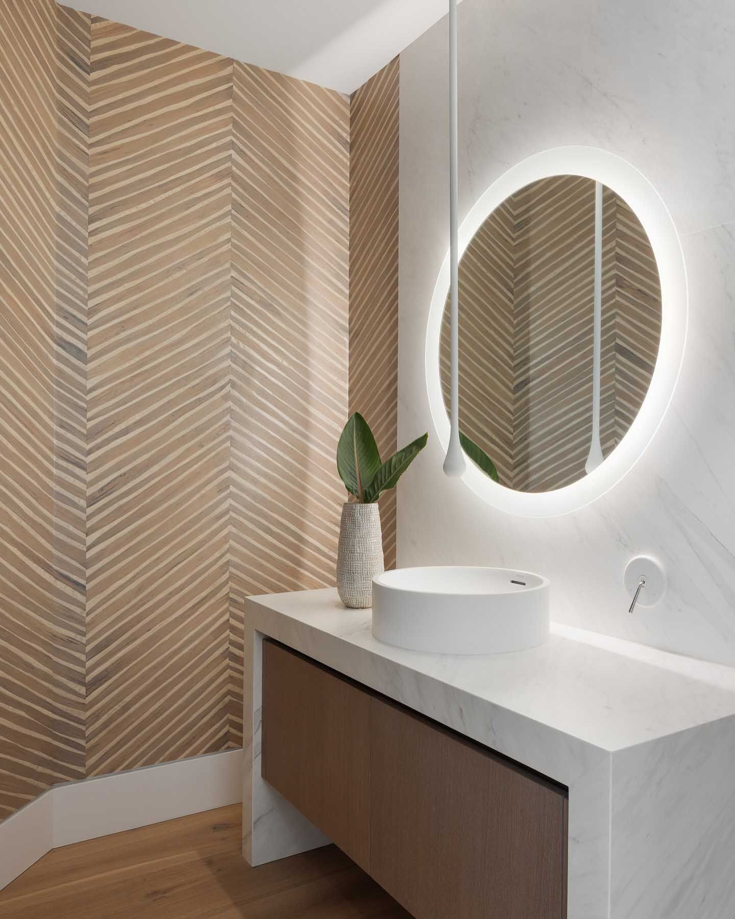 Bathroom with a round, backlit mirror, a vessel sink, and beige, herringbone patterned wallpaper.