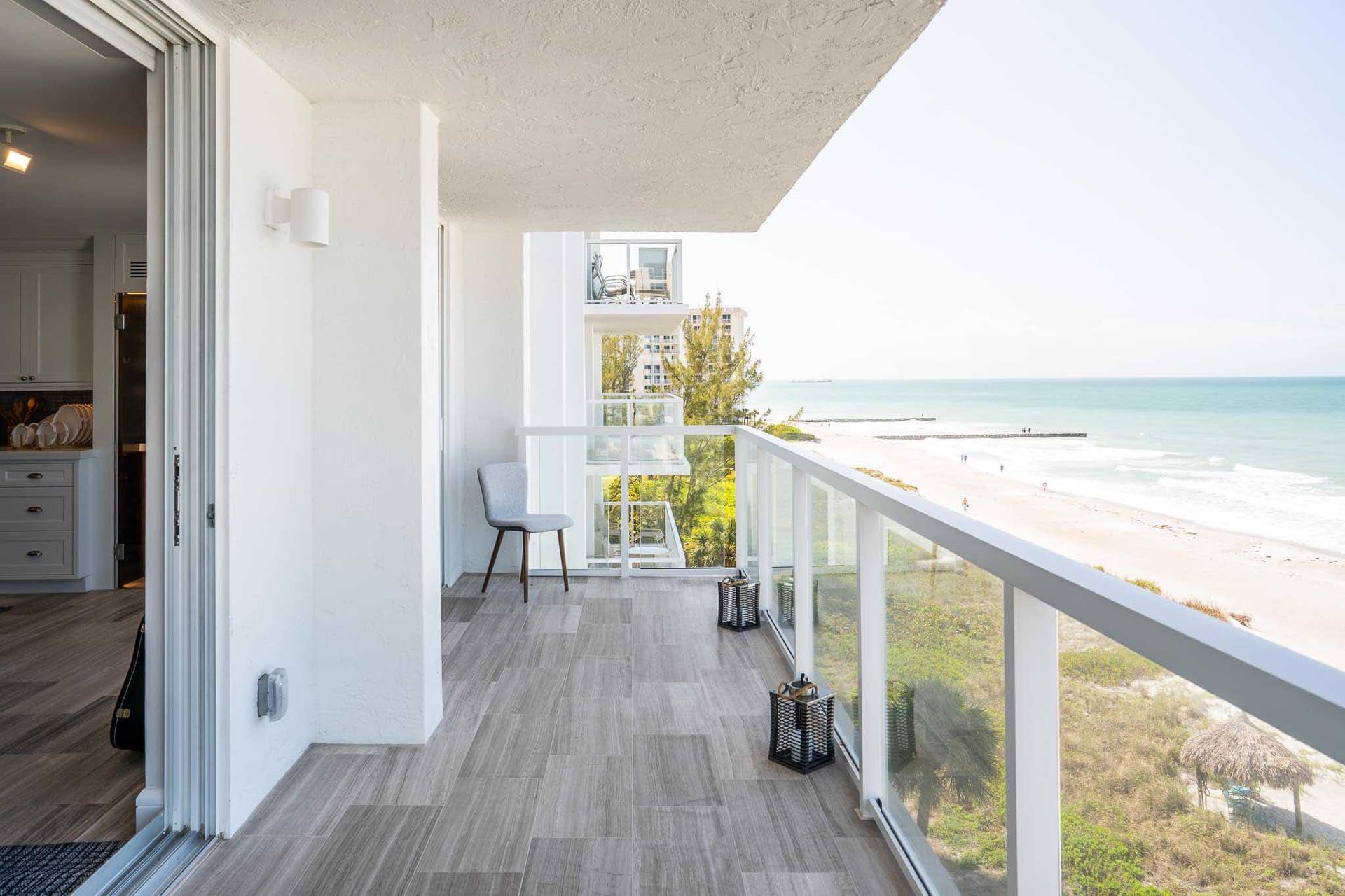 Balcony overlooking a beach, glass railing, gray tile floor, white walls. Ocean view, sunny day.