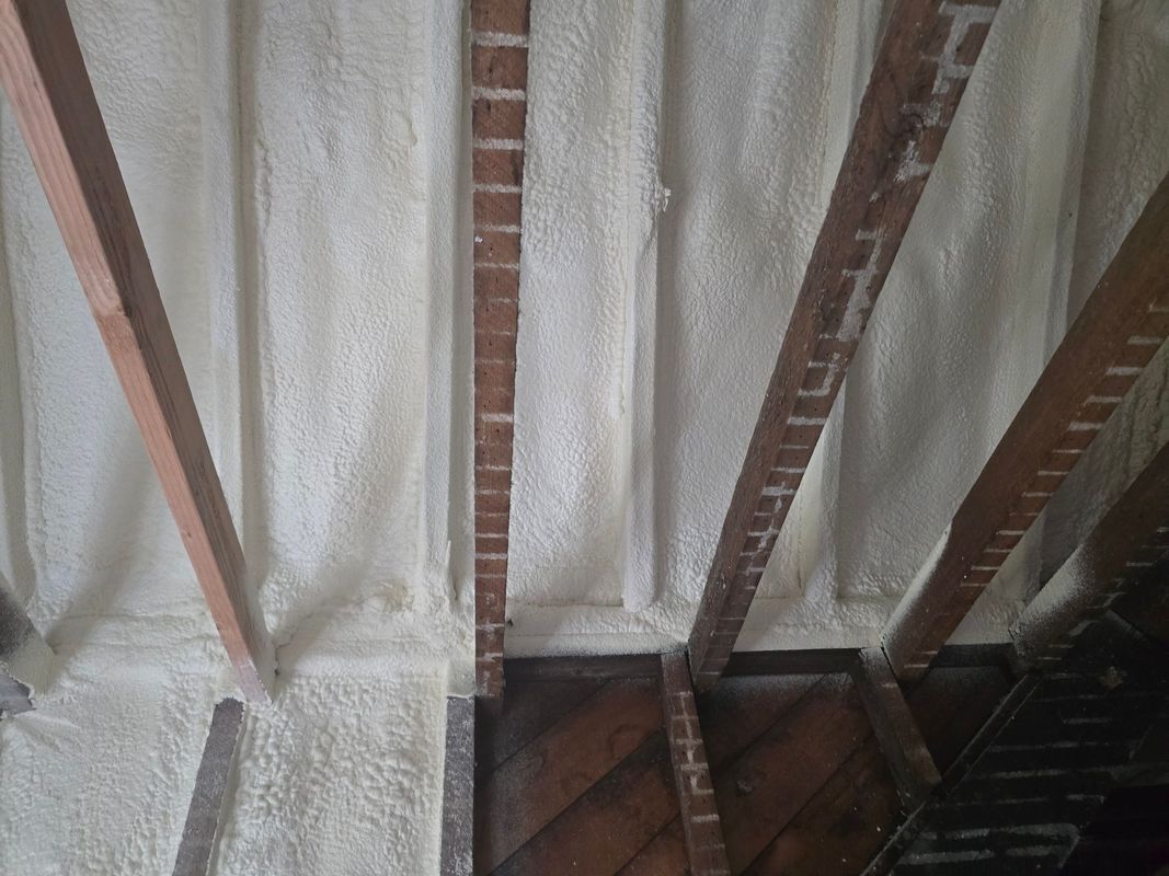 Attic ceiling insulated with light-colored spray foam, covering wood beams and brick.