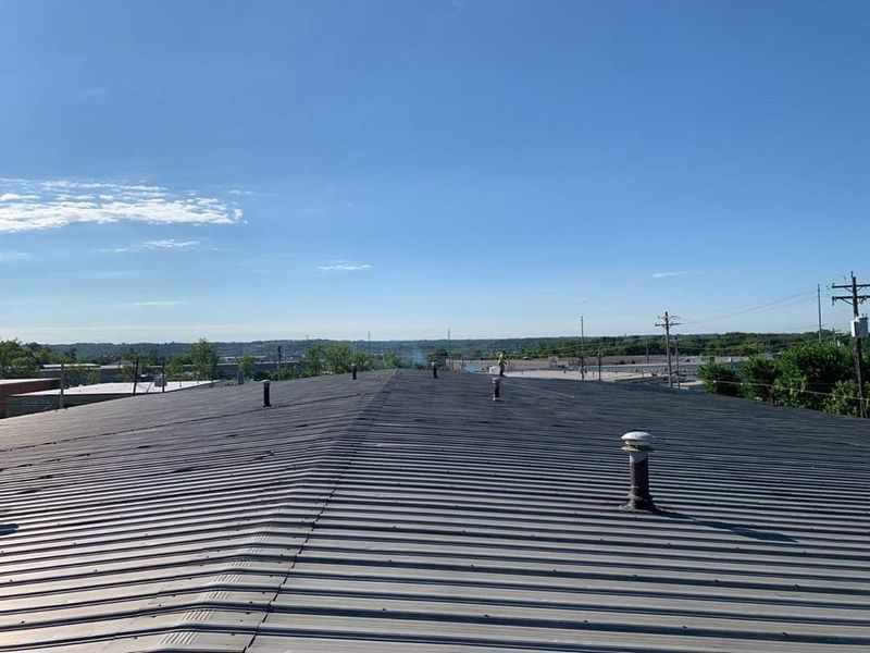 View of a flat, corrugated metal roof under a bright blue sky with sparse greenery visible in the distance.