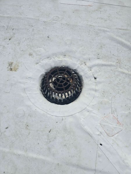 A close-up of a rooftop drain with a metal grate, centered on a white membrane roof.