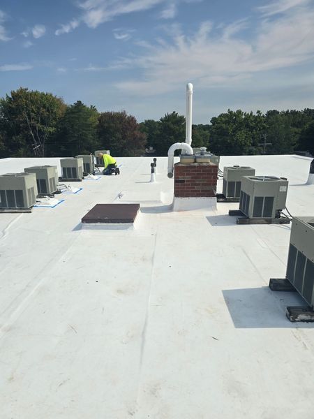 White flat roof with HVAC units, chimney, and a worker in safety gear. Blue sky and trees in the background.