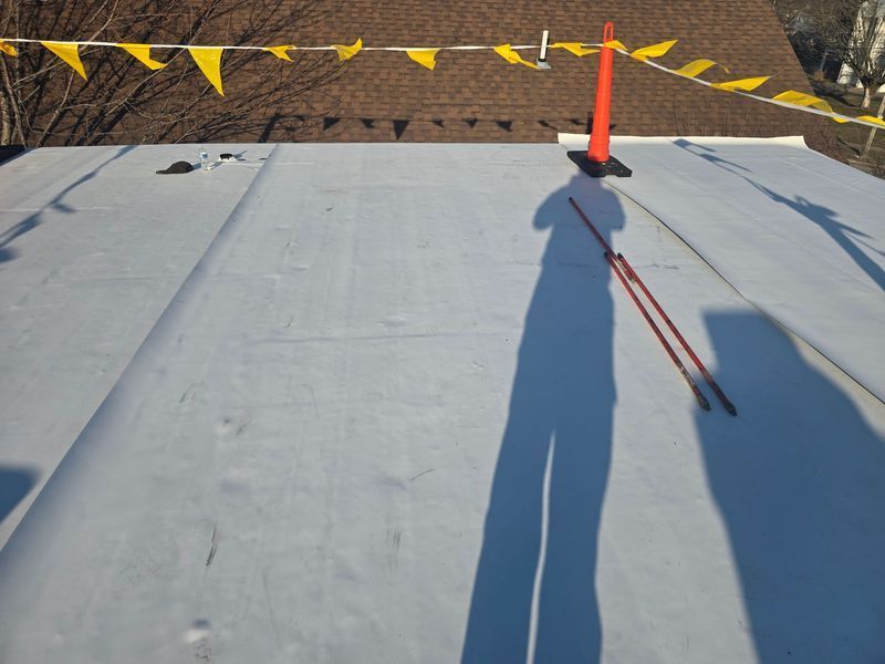 Flat white roof with orange cone, flags, and long shadow of a person holding red tools.