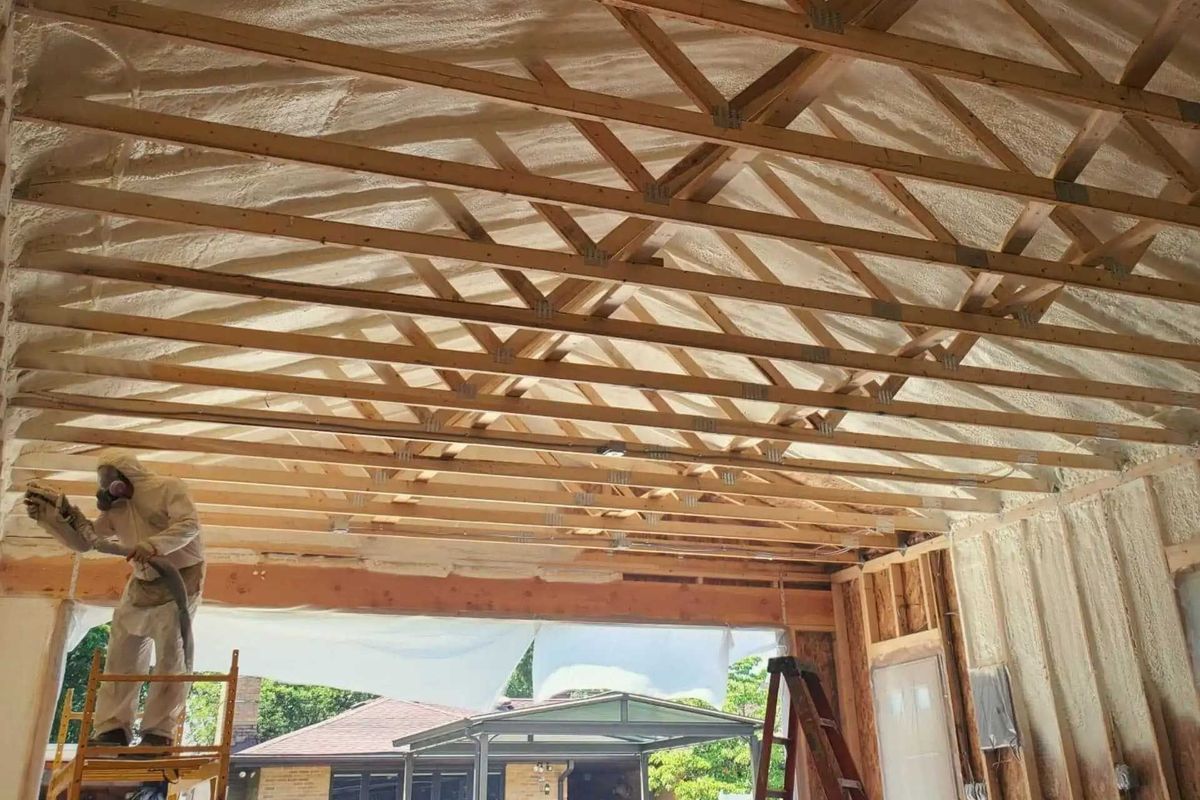 Man in protective suit spraying insulation foam on a wooden ceiling frame.