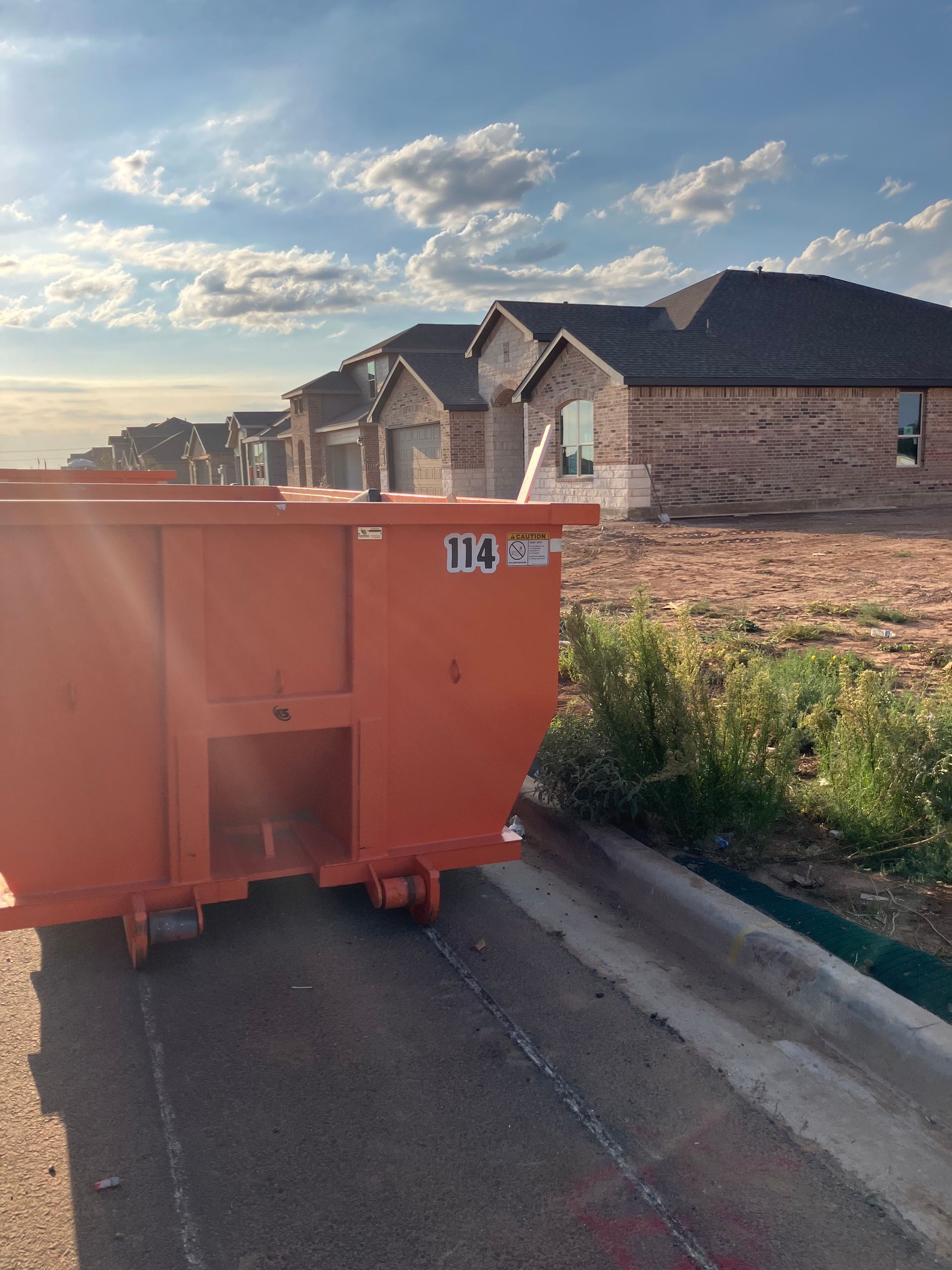 A bright orange construction dumpster sits on an asphalt street in front of a row of newly built houses under a blue sky.