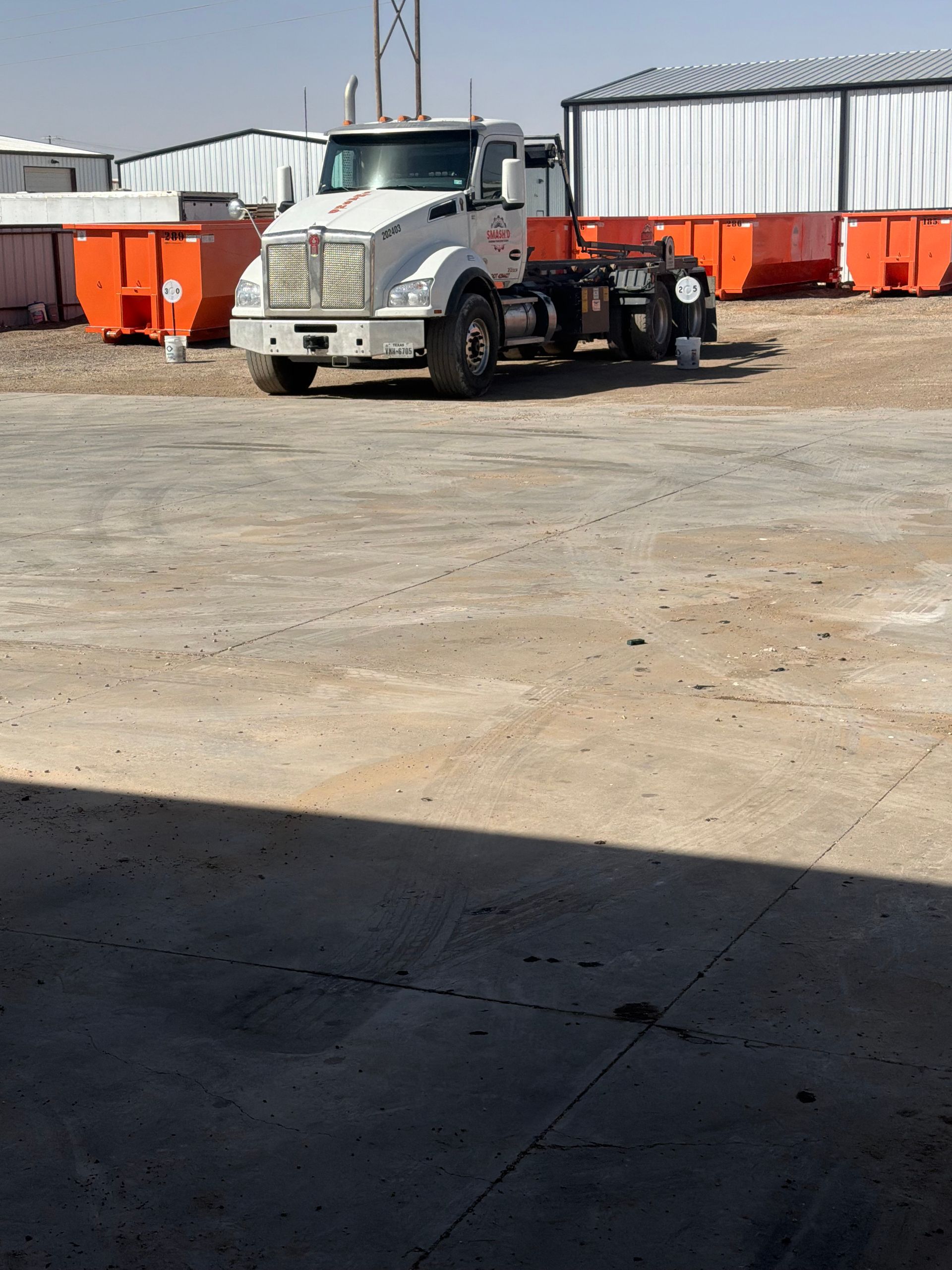 A white semi-truck parked in a gravel lot next to several bright orange dumpsters under a clear sky.
