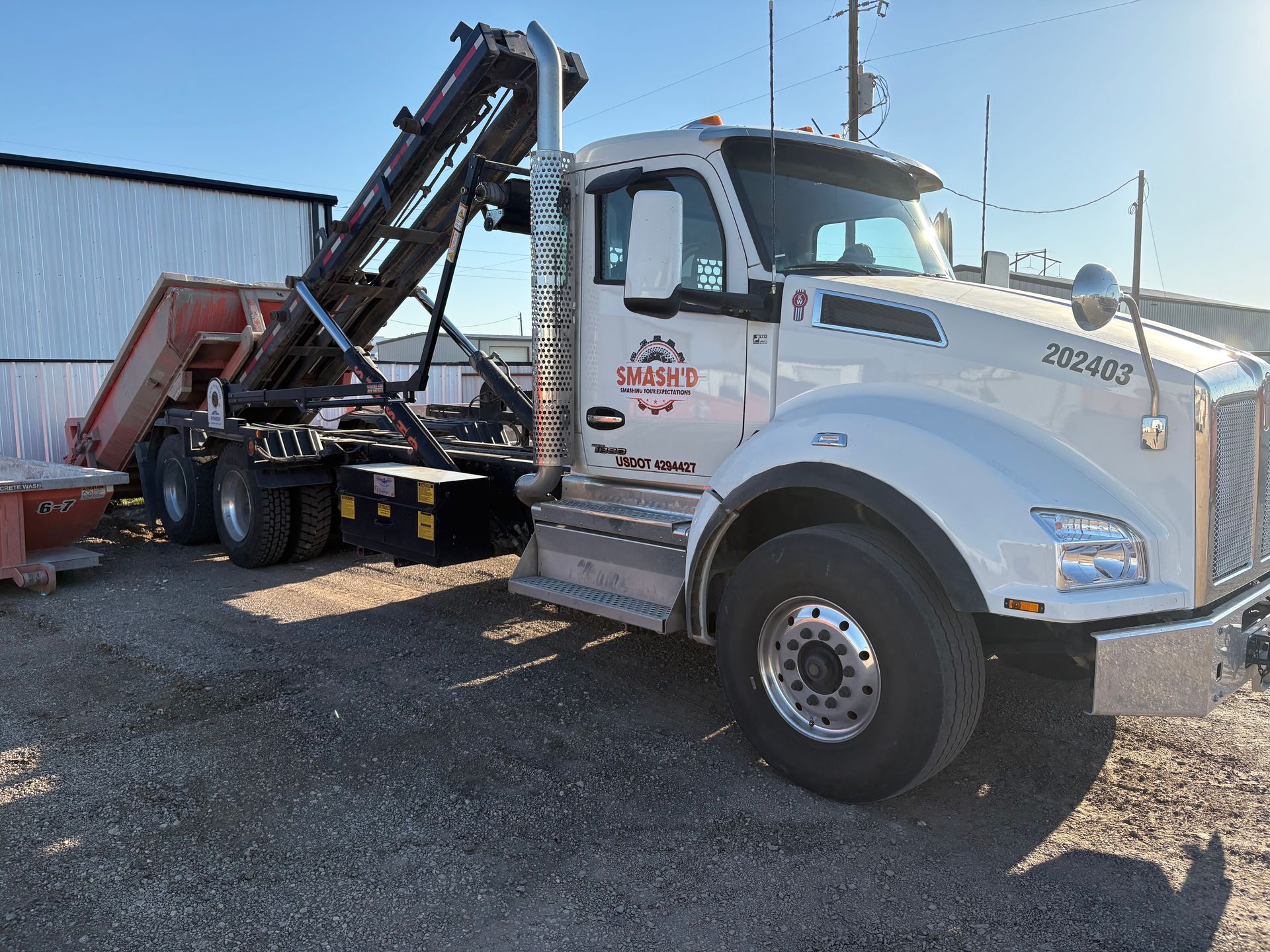 A white roll-off dump truck with a raised hydraulic arm sits on a gravel lot next to a white building.