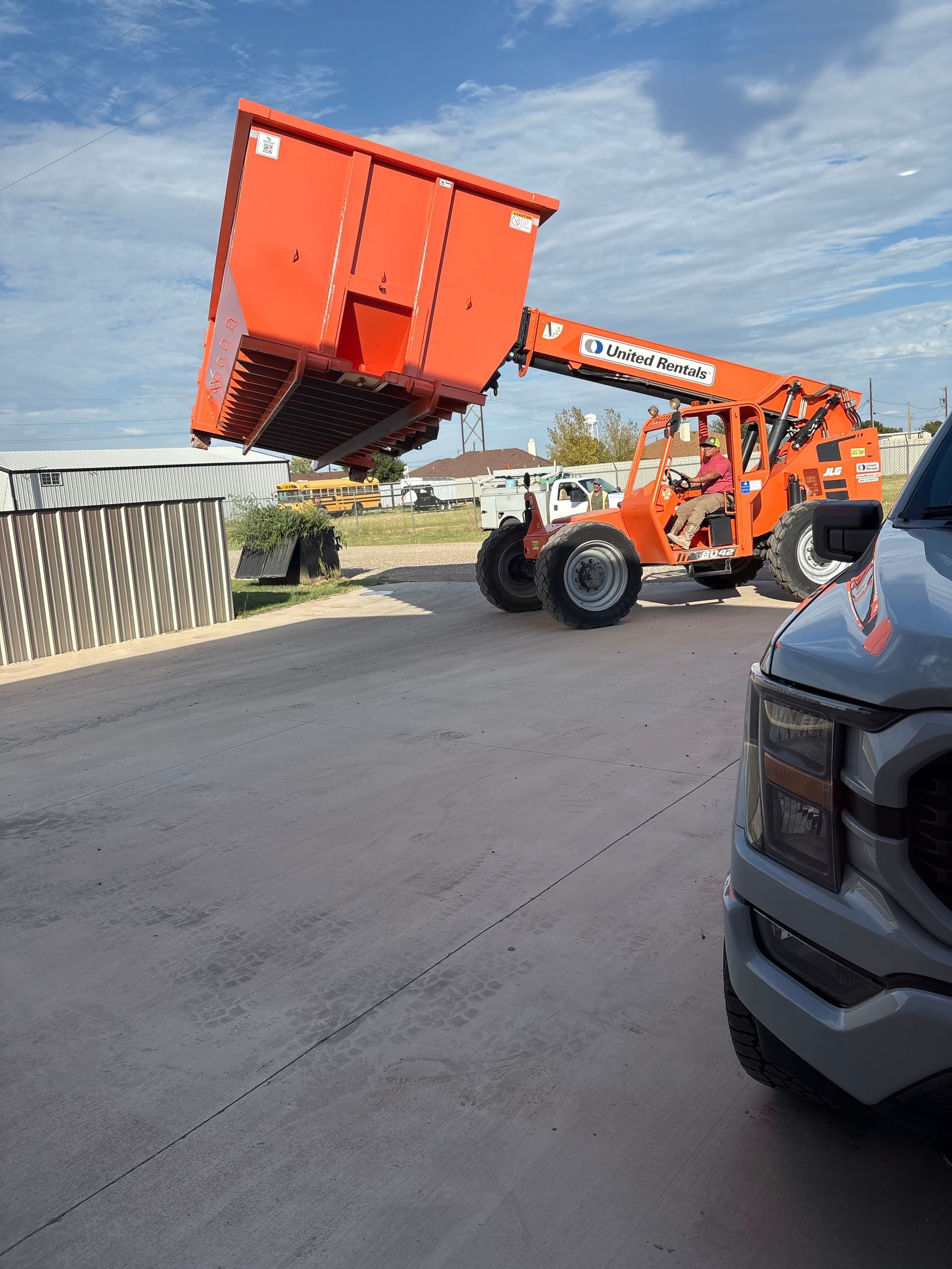 An orange telehandler lifts a large, bright orange industrial hopper outdoors on a sunny day.