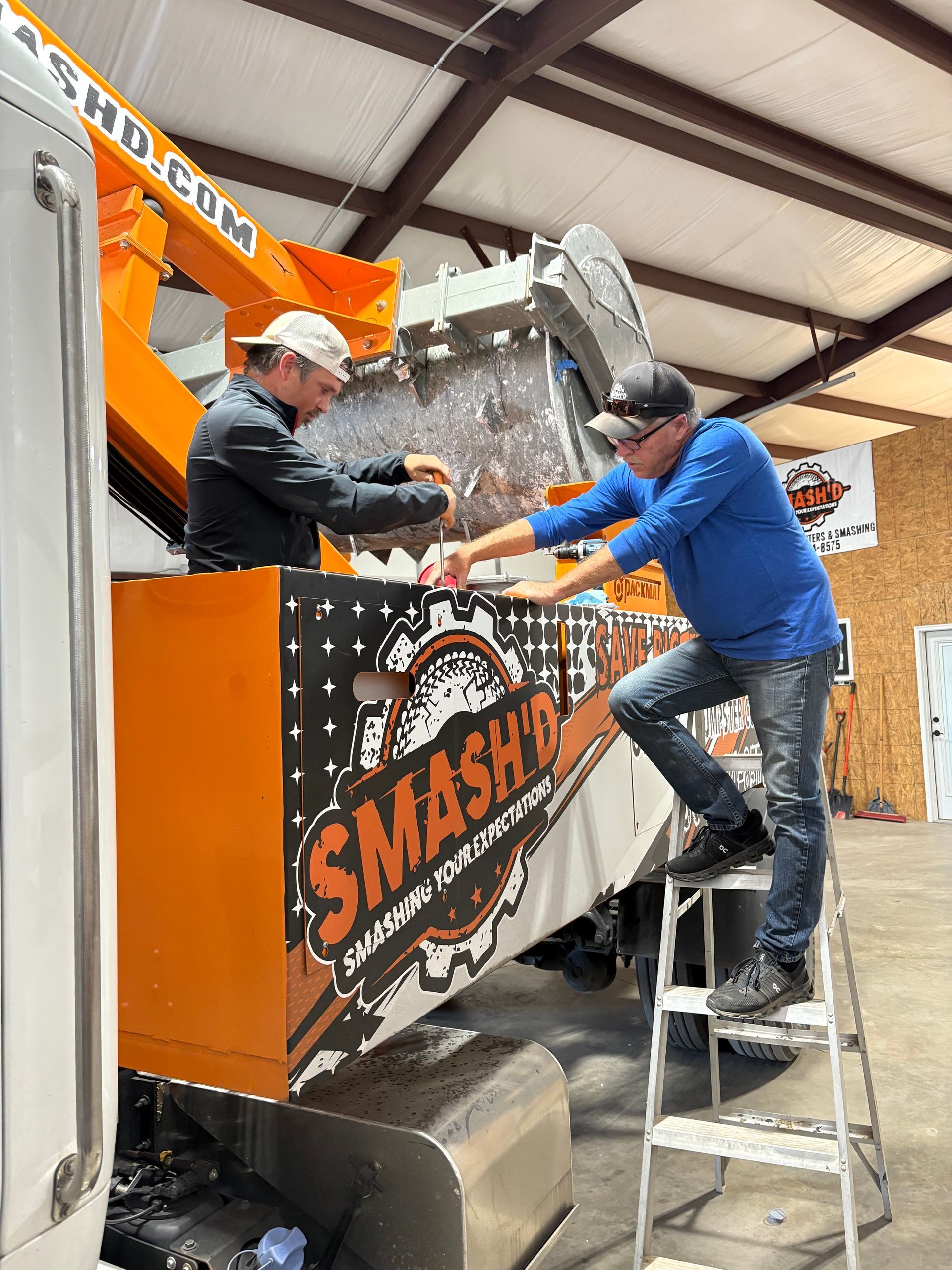 Two people work on a large piece of orange industrial equipment labeled 