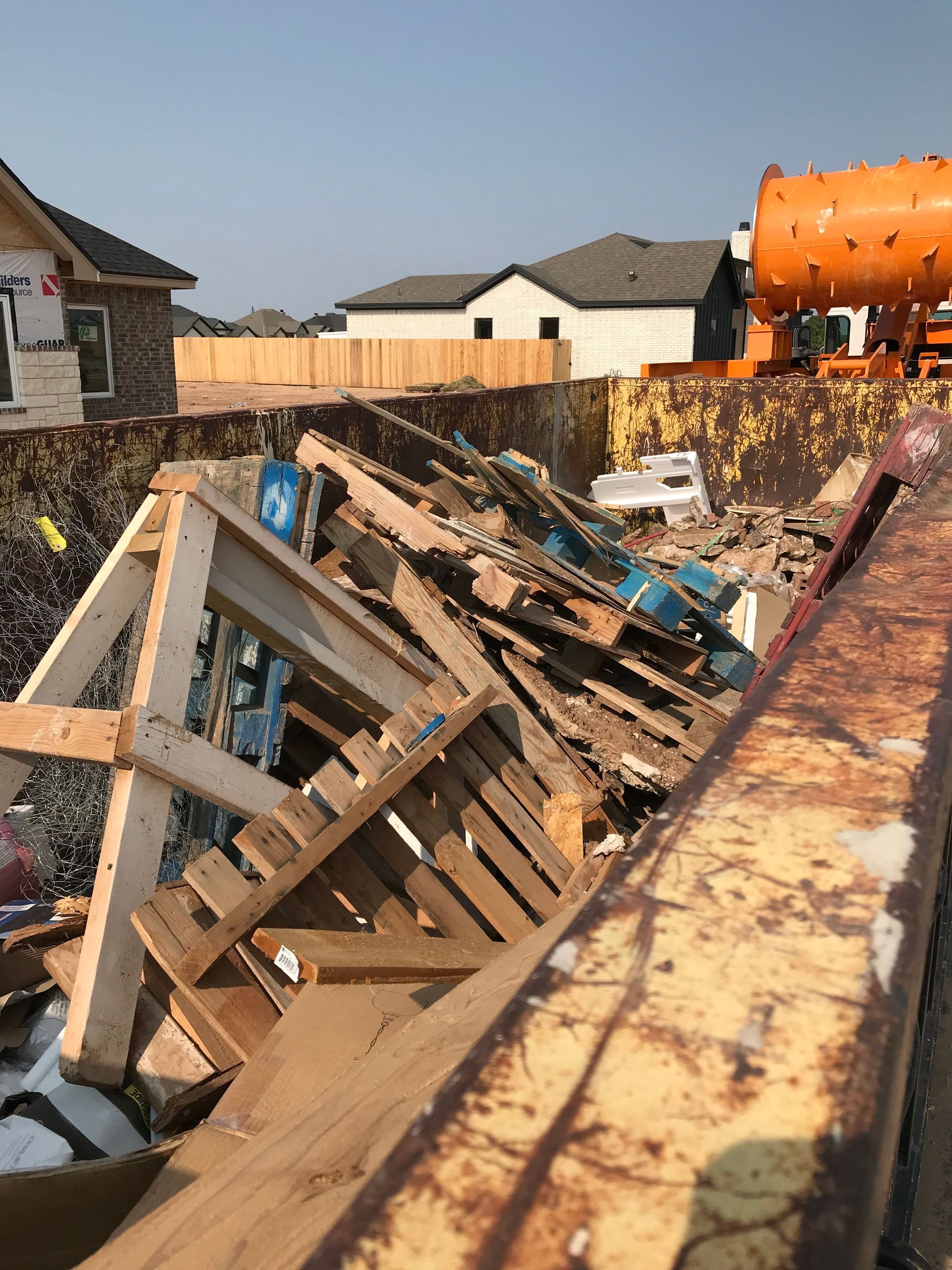 A metal construction dumpster filled with wooden pallets and building debris, set against a backdrop of new houses.