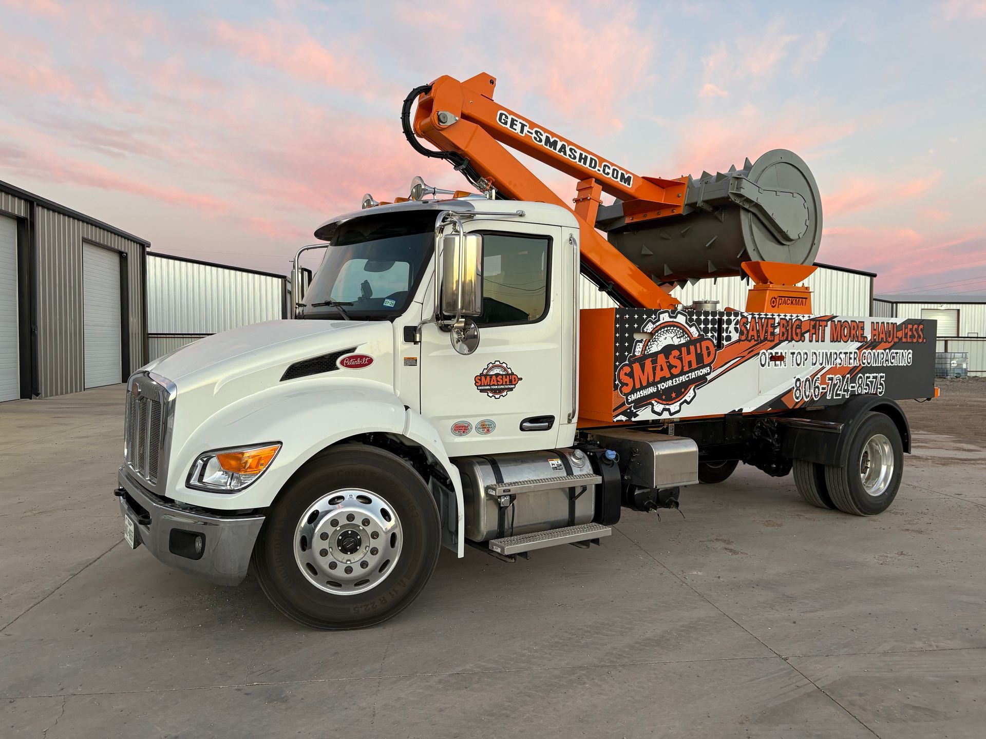 A white Peterbilt truck with an orange boom and hydraulic crushing equipment, parked on a paved lot at sunset.