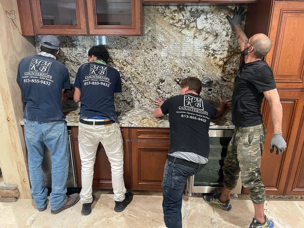 Four workers installing a granite backsplash in a kitchen with brown cabinets and a small wine fridge.