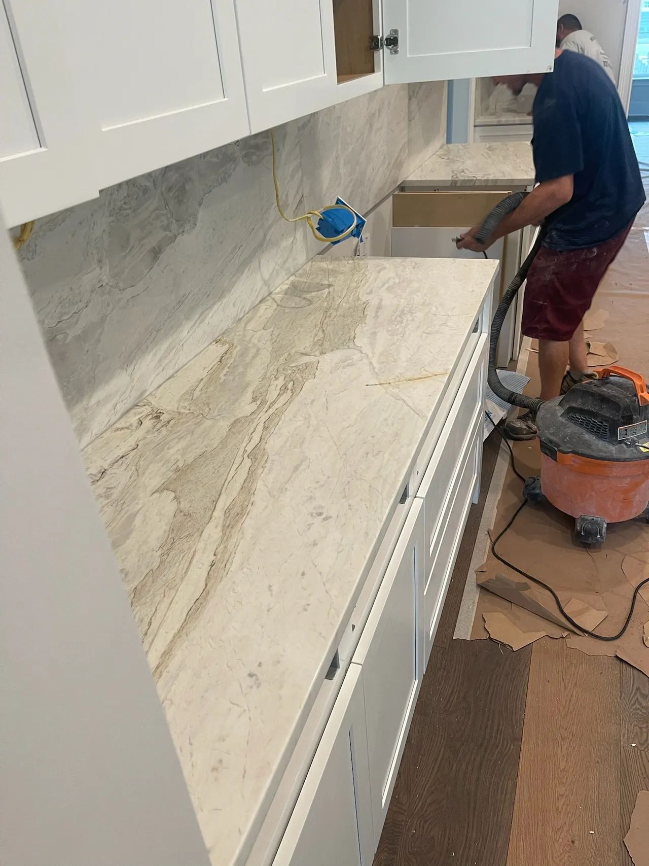 Person vacuuming a kitchen countertop with light-colored stone. White cabinets and backsplash are visible.