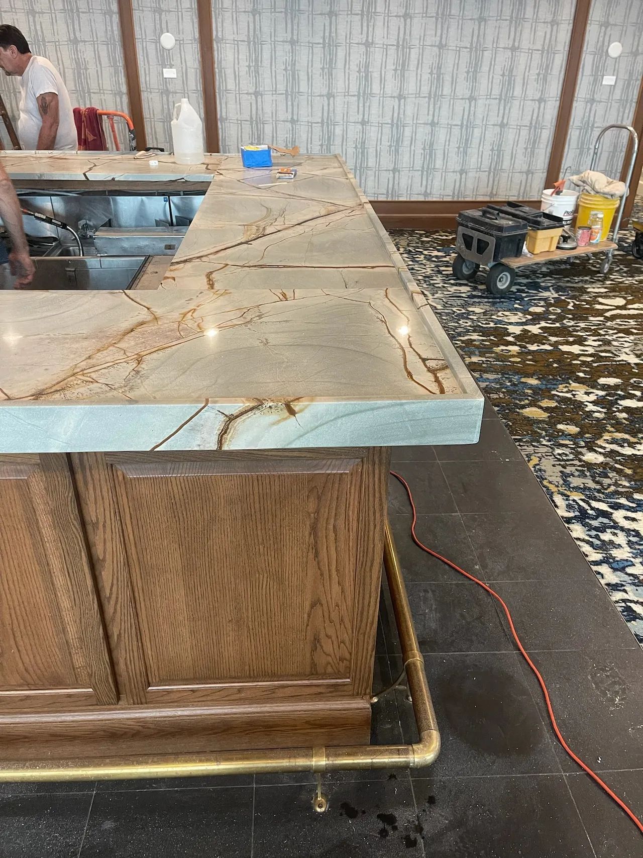 A bar with a stone countertop, wood cabinetry, and a brass footrail; workers are present in the background.