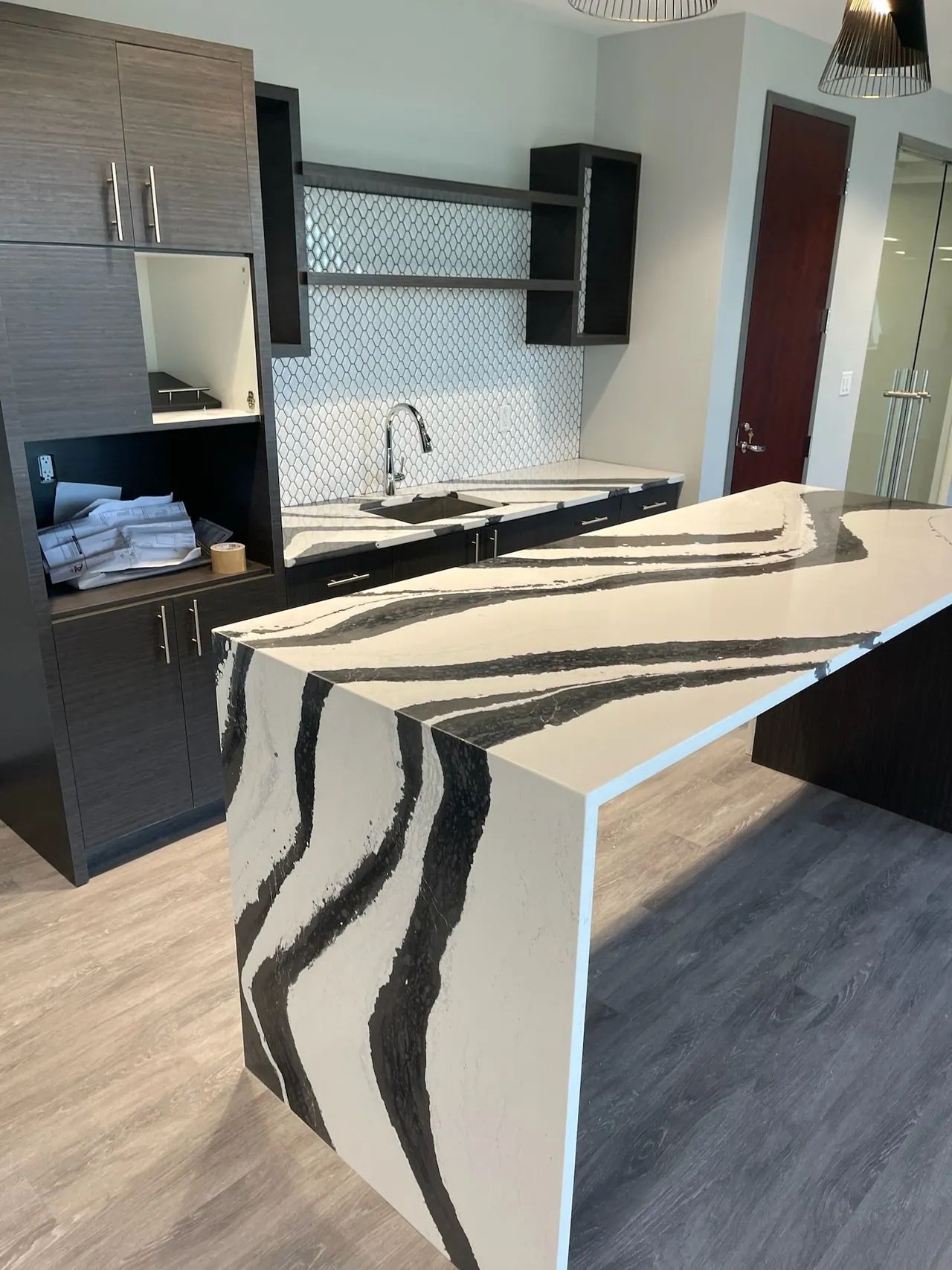 Modern kitchen with white and black patterned countertop, dark cabinets, and stainless steel backsplash.