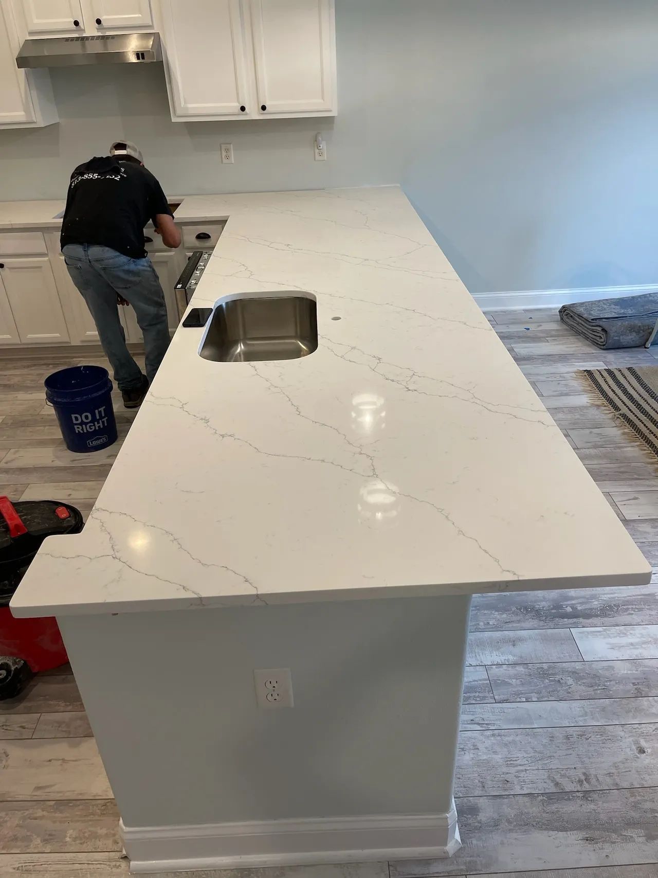 A person works on a white countertop island with a sink in a kitchen with white cabinets and gray flooring.