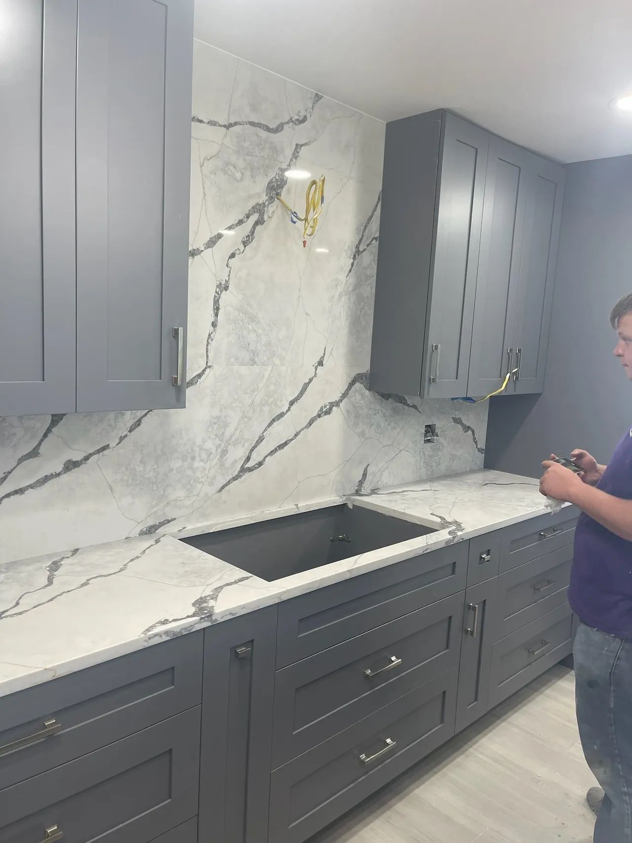 Gray kitchen with marble backsplash, cabinets, and countertops. Man standing on the right.