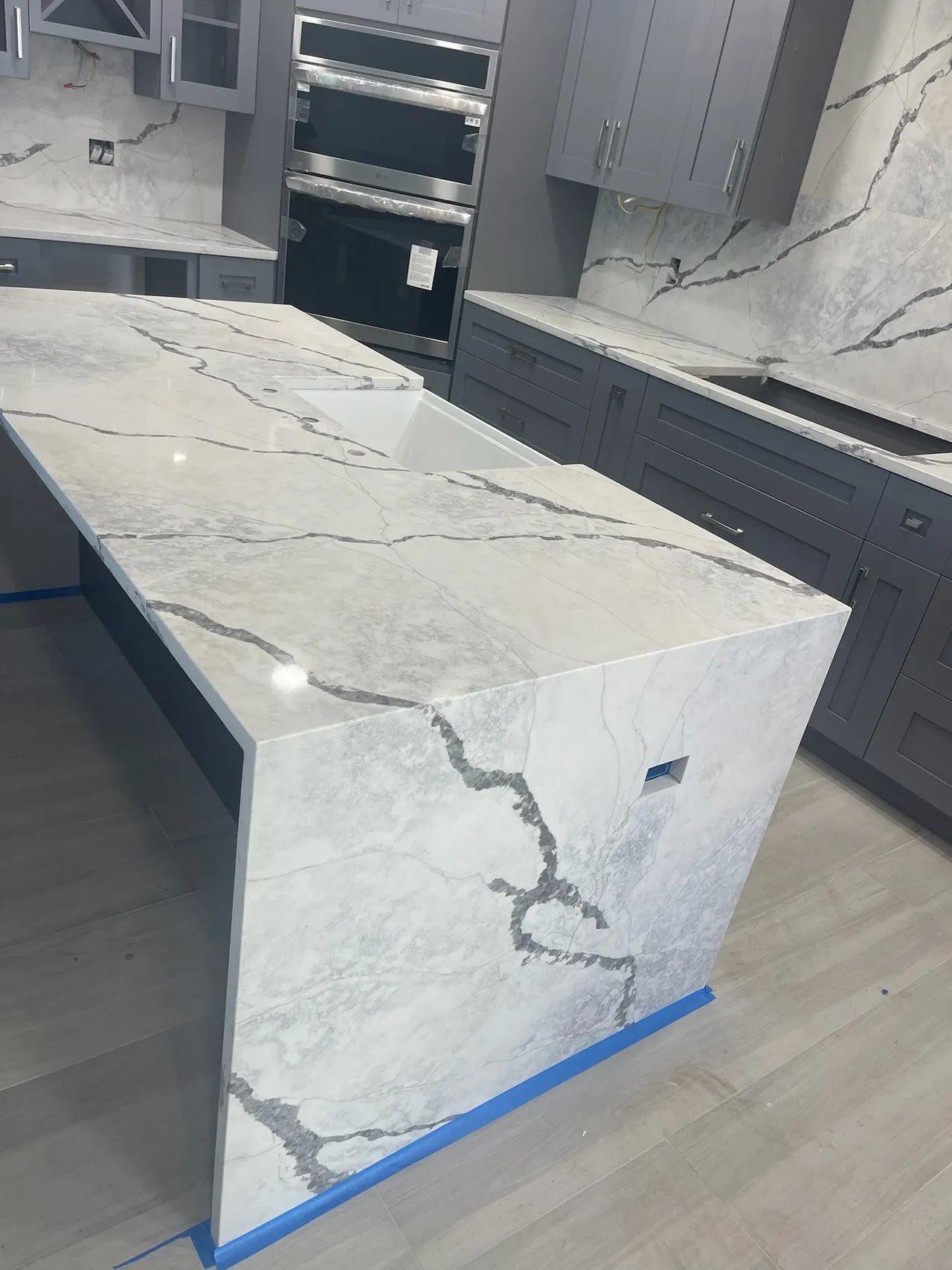 A modern kitchen with a marble island countertop, grey cabinets, and built-in appliances.