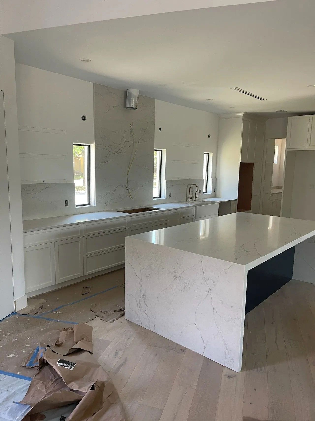 Kitchen under construction with white cabinets, island, and countertops. Unfinished walls, wood floors.