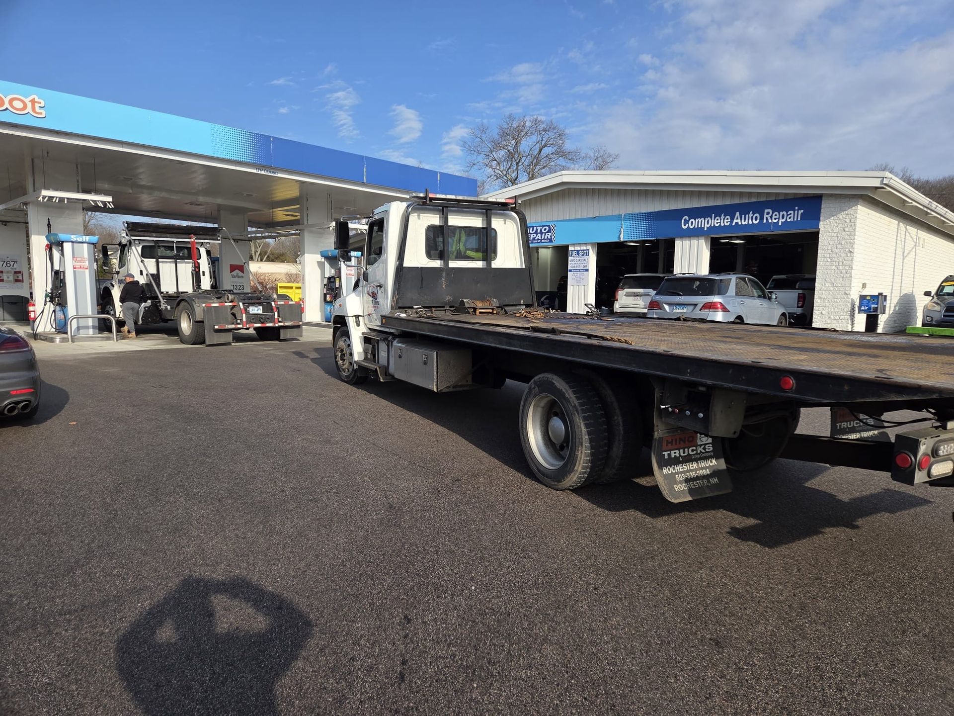 Flatbed tow truck at a gas station, with a car nearby under a blue canopy.