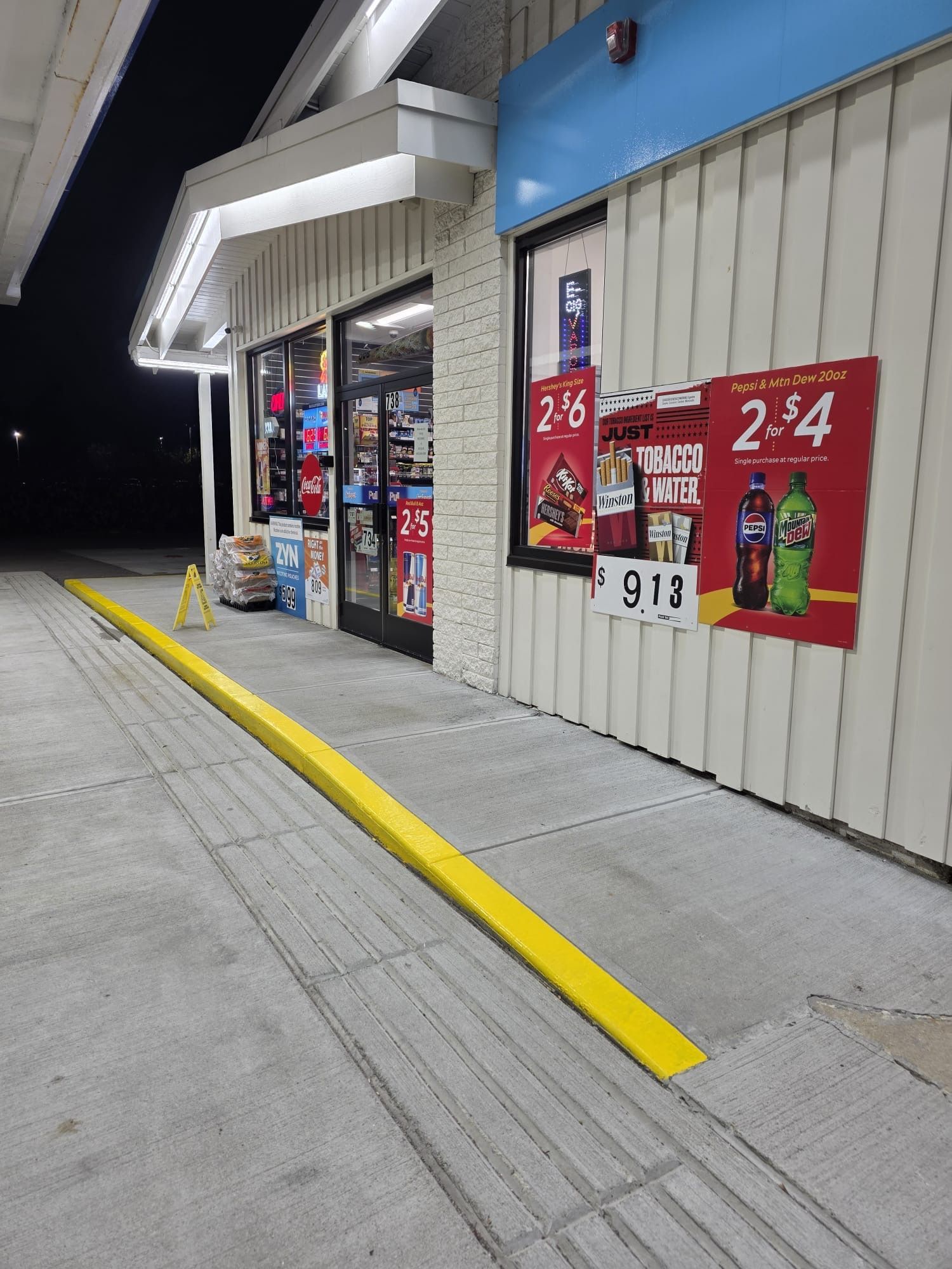 Convenience store exterior at night with bright red sale signs and a yellow curb ramp by the entrance
