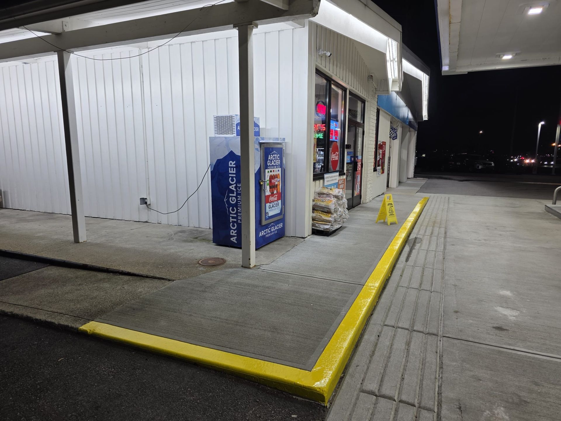 Corner of a convenience store at night with bright yellow curb and vending machines by the entrance