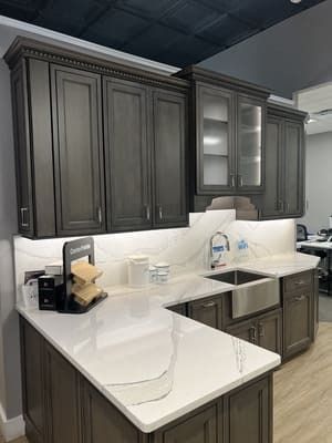 A kitchen with gray cabinets , white counter tops , and a sink.