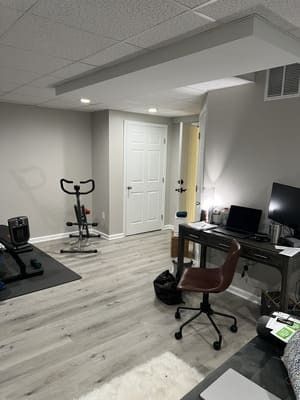 A basement with a desk , chair , laptop and exercise bike.