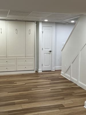 A basement with hardwood floors , white cabinets , and stairs.