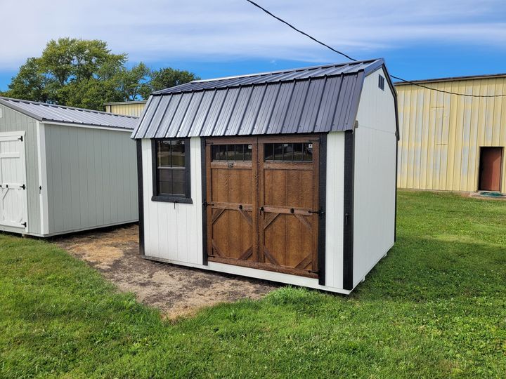A white shed with a black roof and wooden doors is sitting in a grassy field.