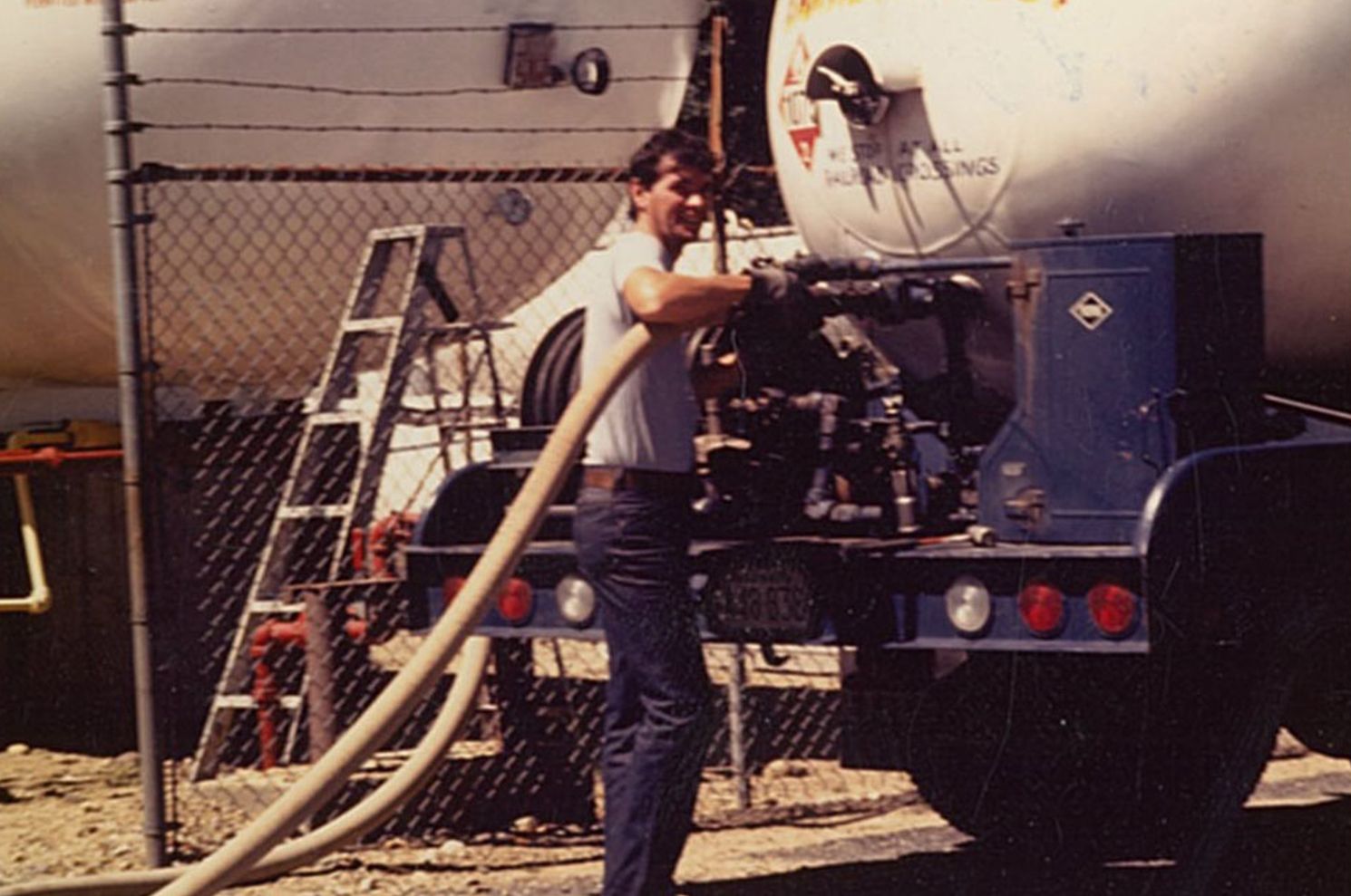 Man in jeans and t-shirt filling a tank with a hose. Tanker truck in a yard near a chain-link fence.