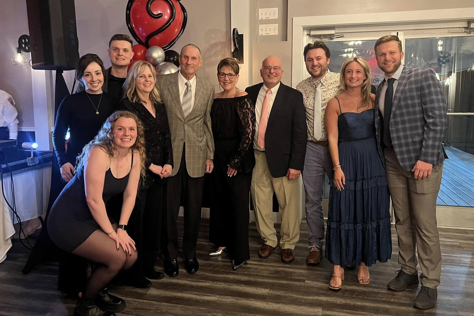 Group of people smiling at a party; woman in blue dress, man in plaid jacket, balloons in background.