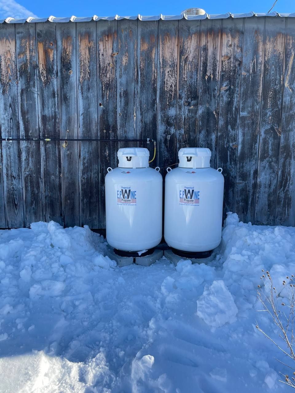 Two white propane tanks in snow against a weathered wooden wall.