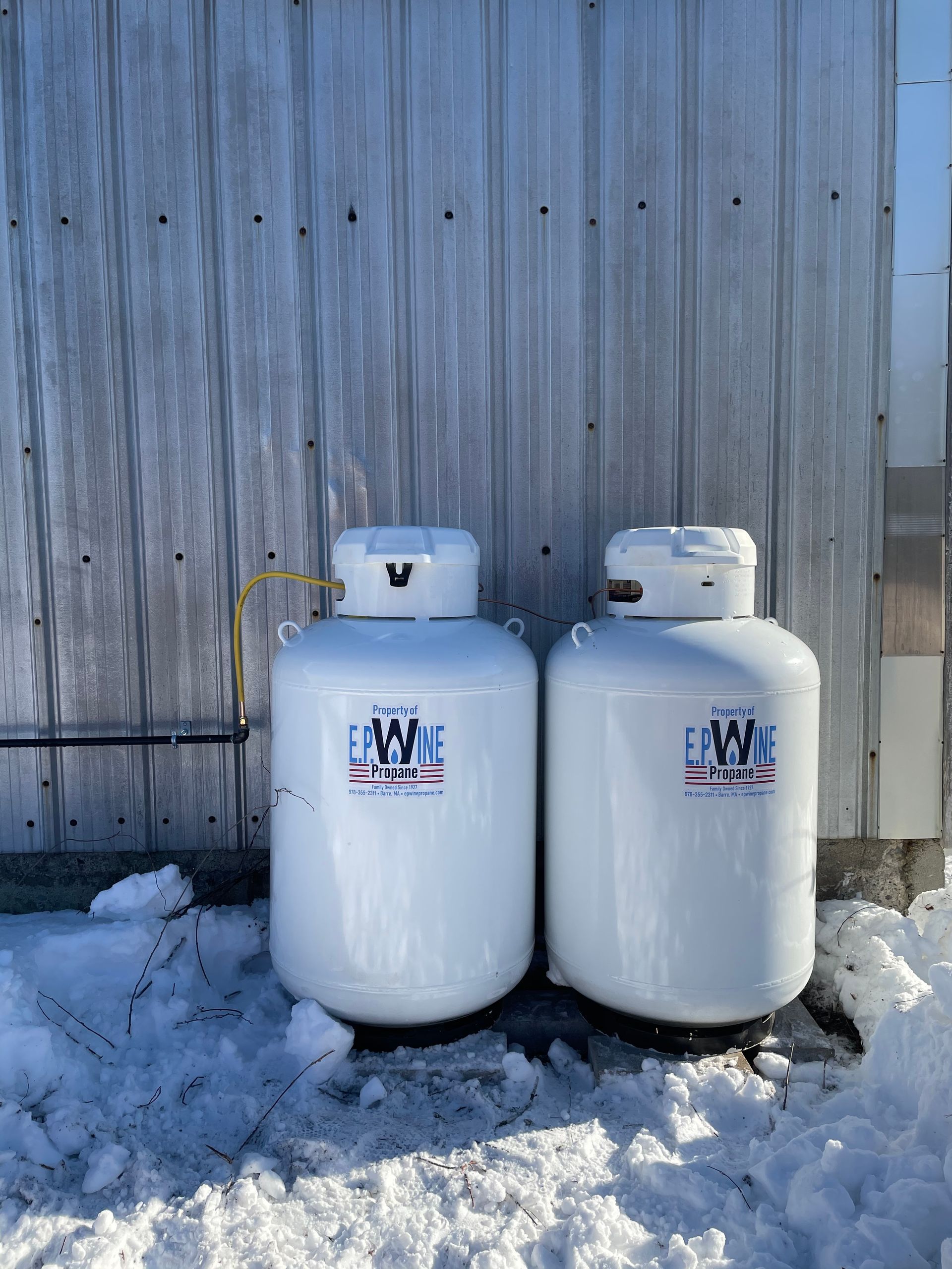 Two white propane tanks against a corrugated metal wall, in a snow-covered outdoor setting.