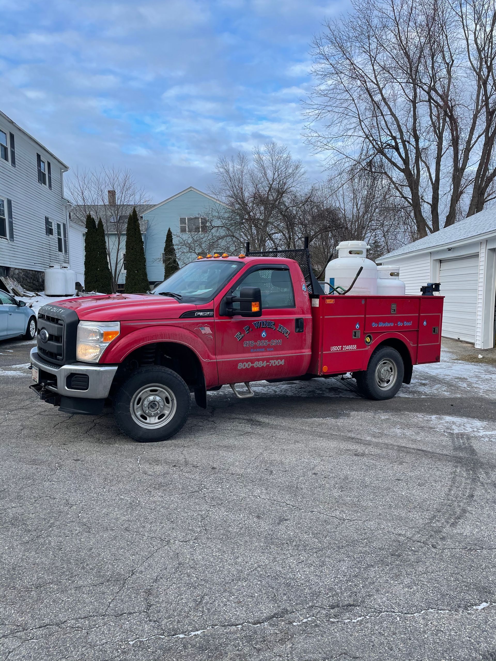 Red work truck parked in a paved lot. Houses and a garage are in the background.