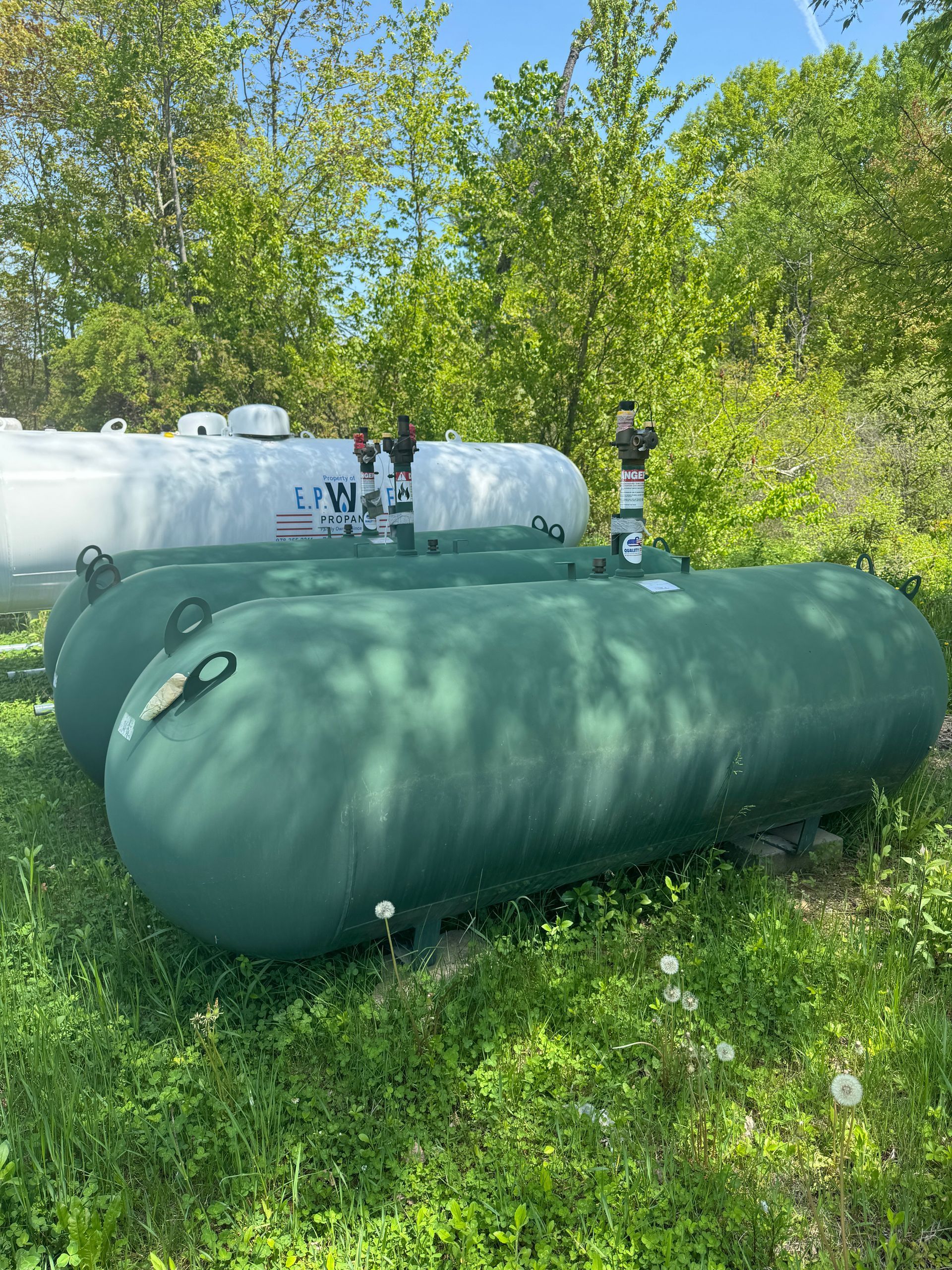 Green propane tanks in a grassy area with a white tank in the background.