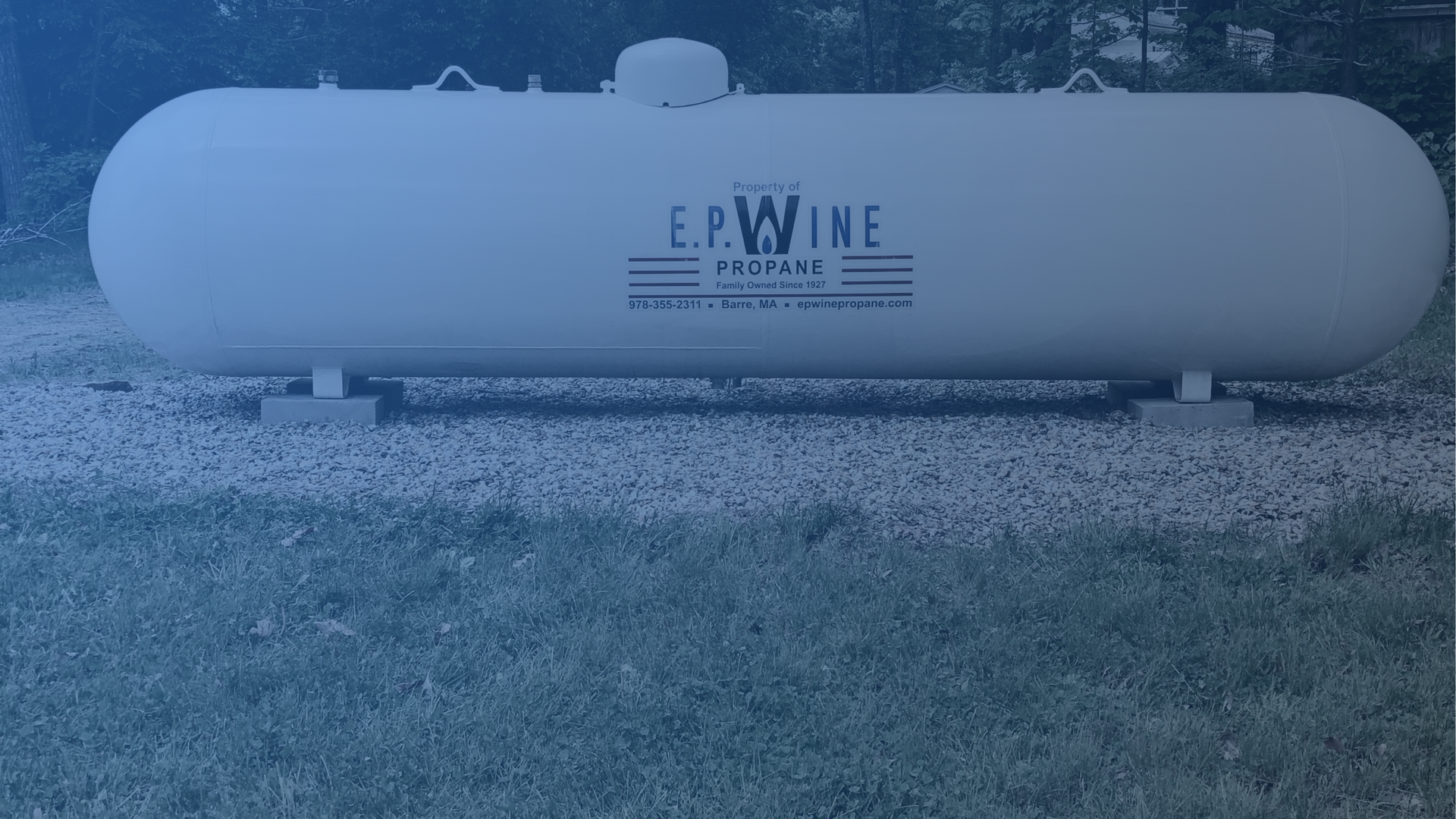 White propane tank on grass in front of trees.