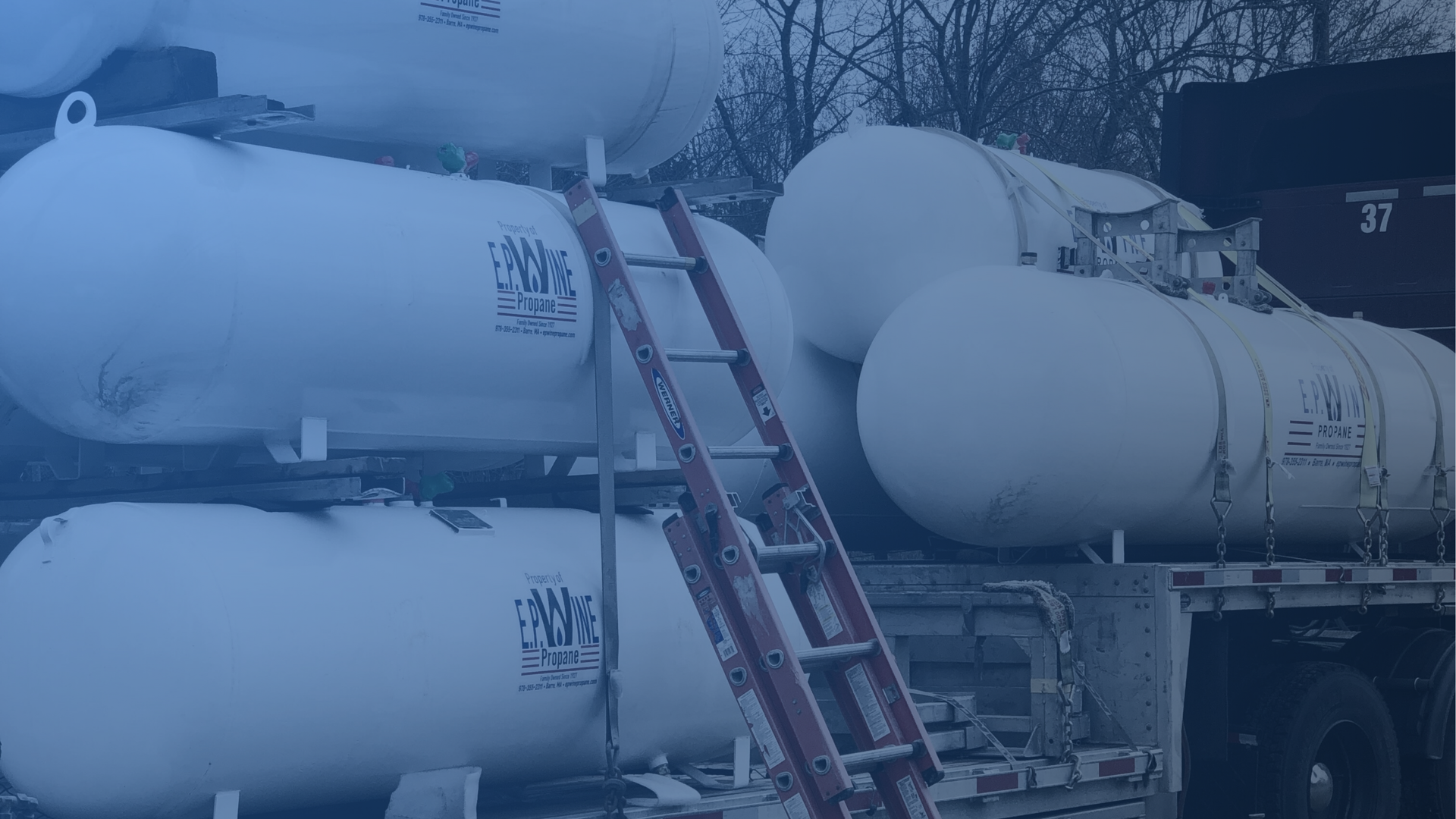 Red truck next to a large white propane tank, likely for delivery. Outdoor setting.