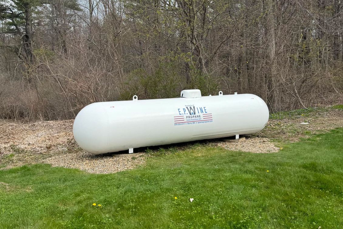 White propane tank on grass in front of trees.