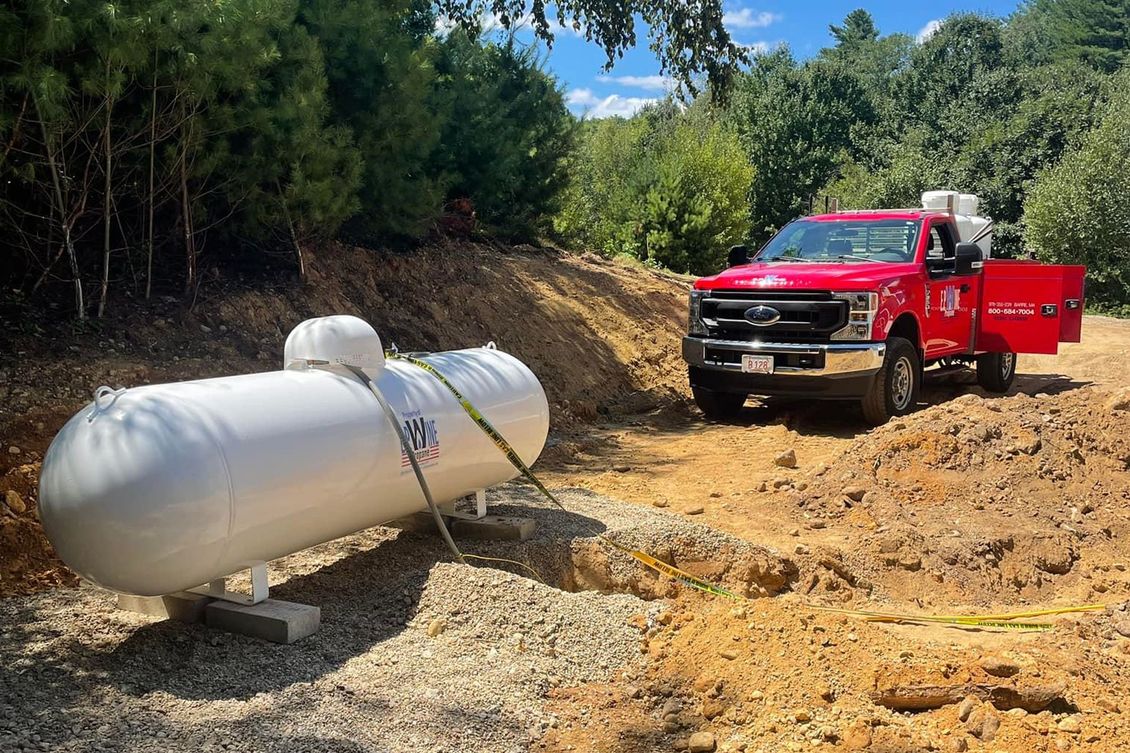Red truck next to a large white propane tank, likely for delivery. Outdoor setting.