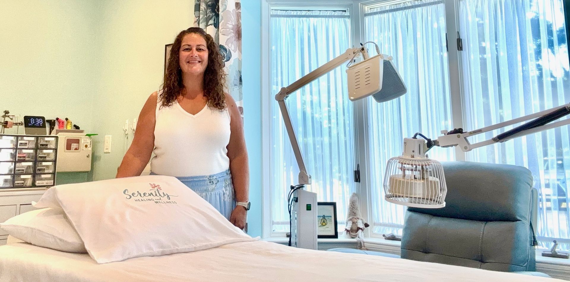 Woman in white top, standing by bed with medical equipment. Window behind her.