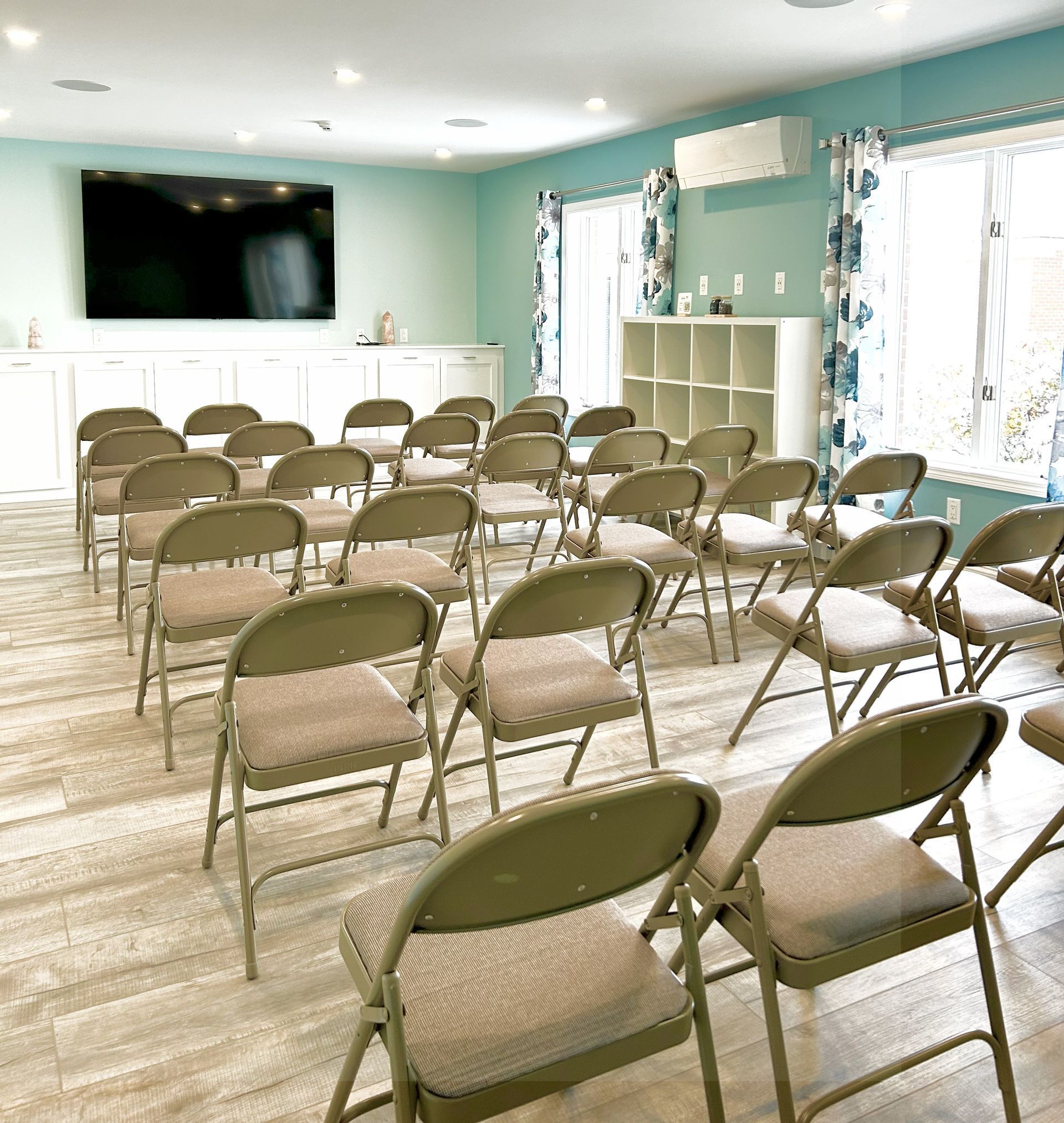 A light-blue room with rows of tan folding chairs facing a large TV; windows with floral curtains on the right.
