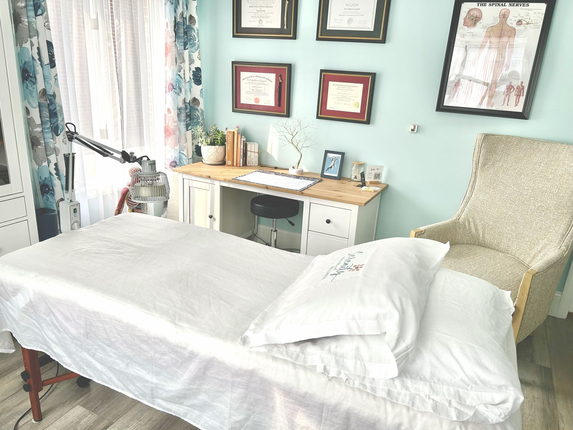 Massage therapy room: white sheets on table, desk with diplomas, light blue walls, beige chair.