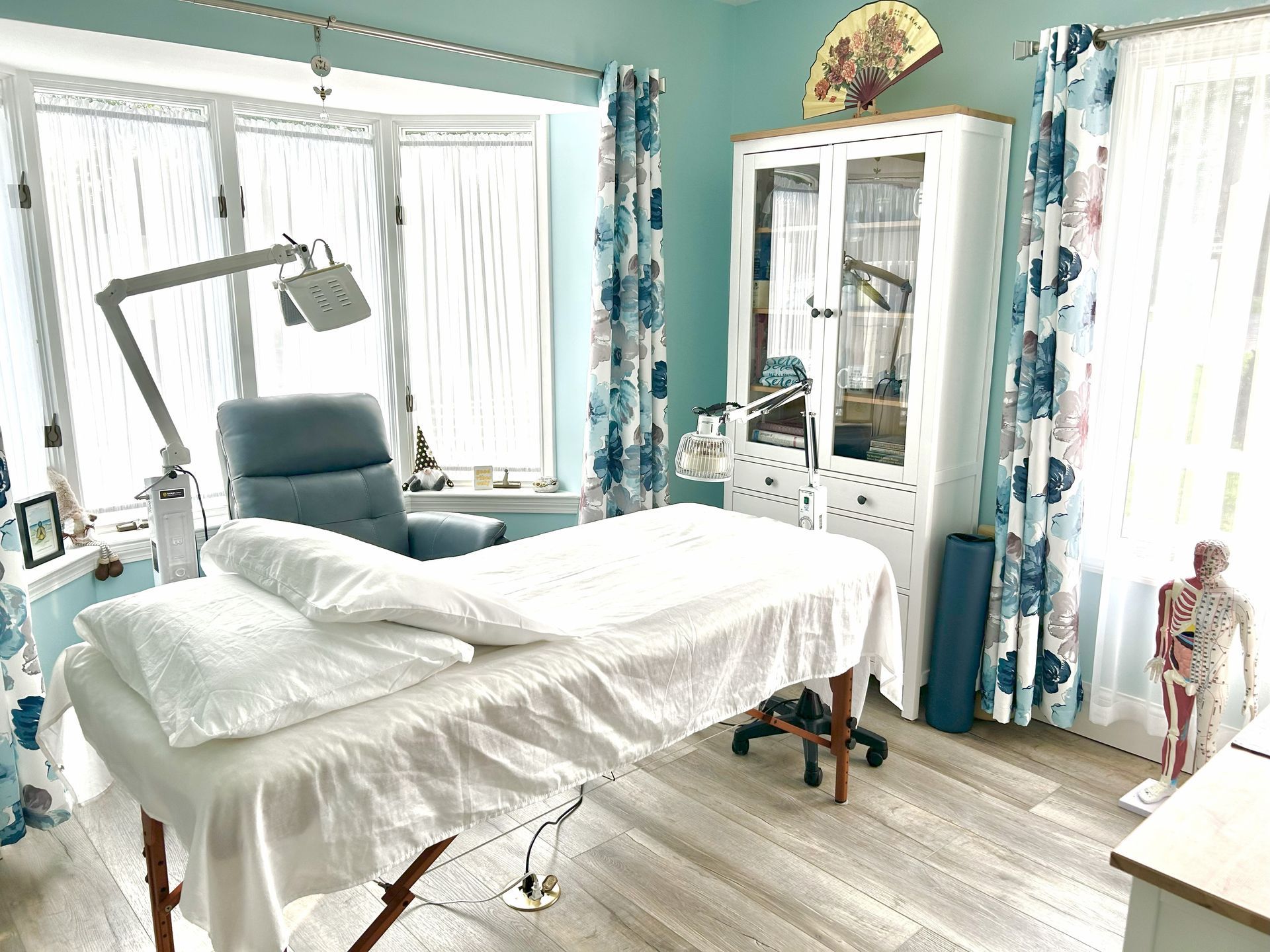 Massage therapy room with a massage table, recliner, and a cabinet next to a window with floral curtains.