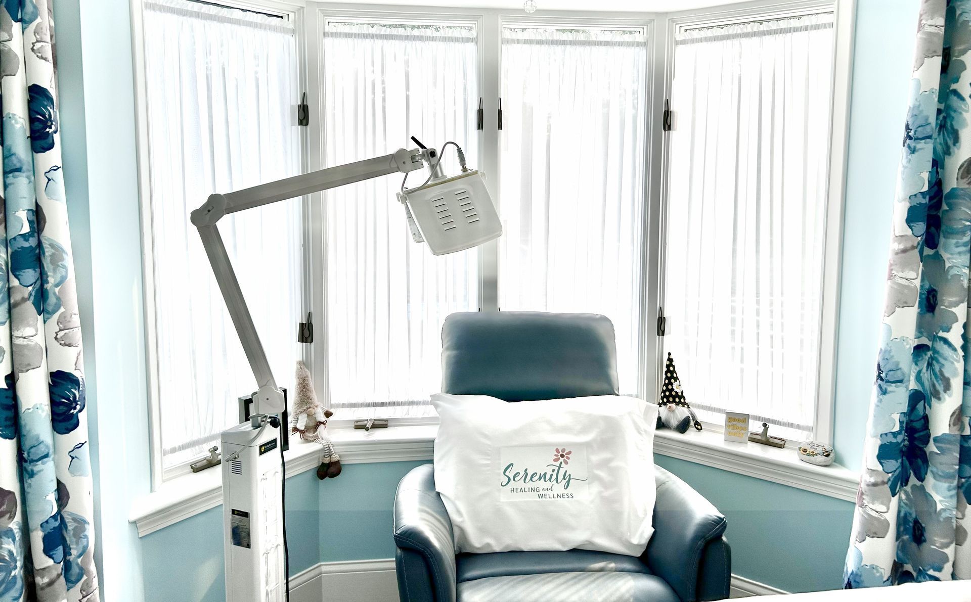 Chair with light therapy equipment in front of a bay window with floral curtains.