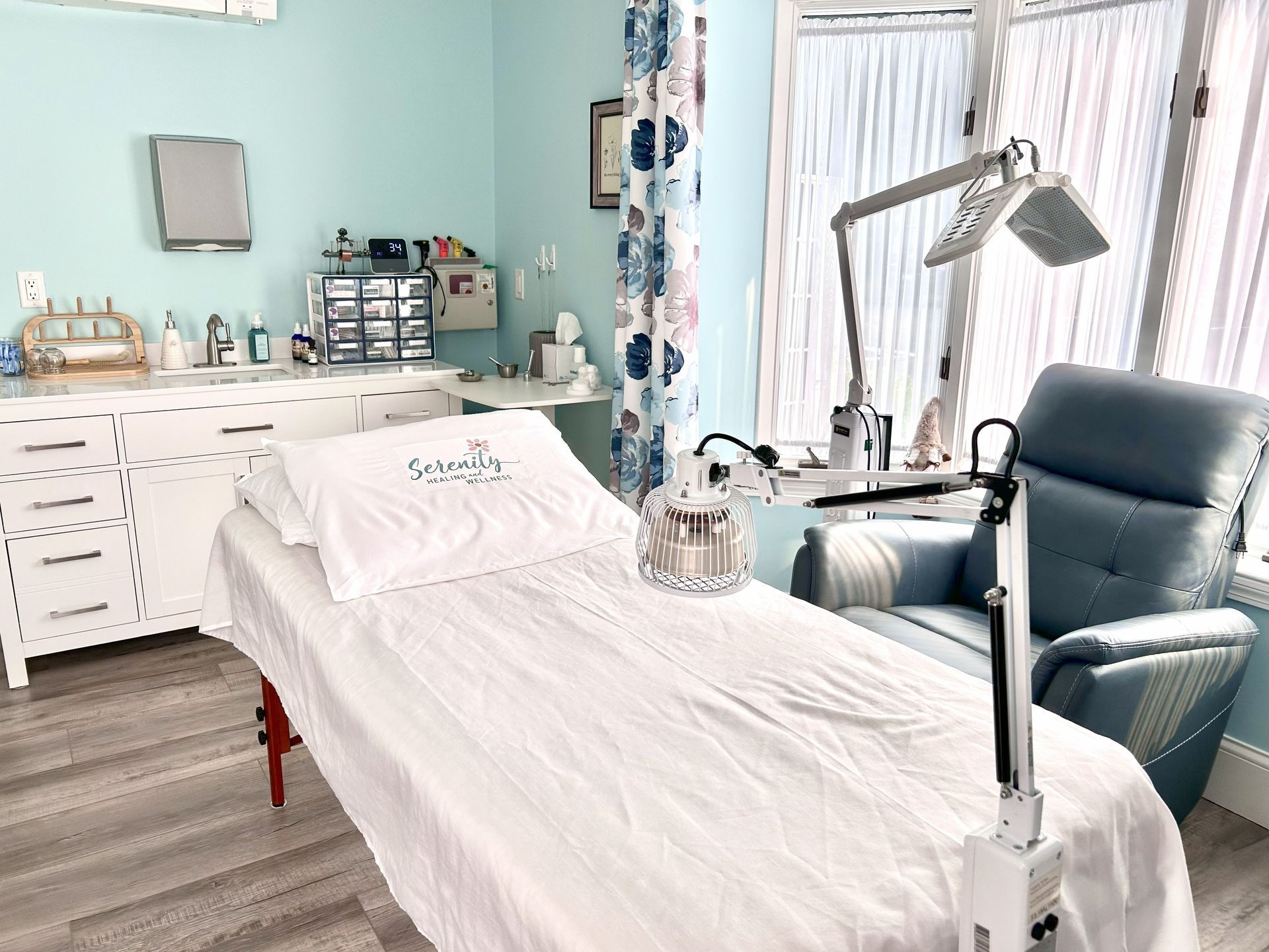 Aesthetician's treatment room: blue walls, white furniture, treatment bed, recliner, light fixture, window with sheers.