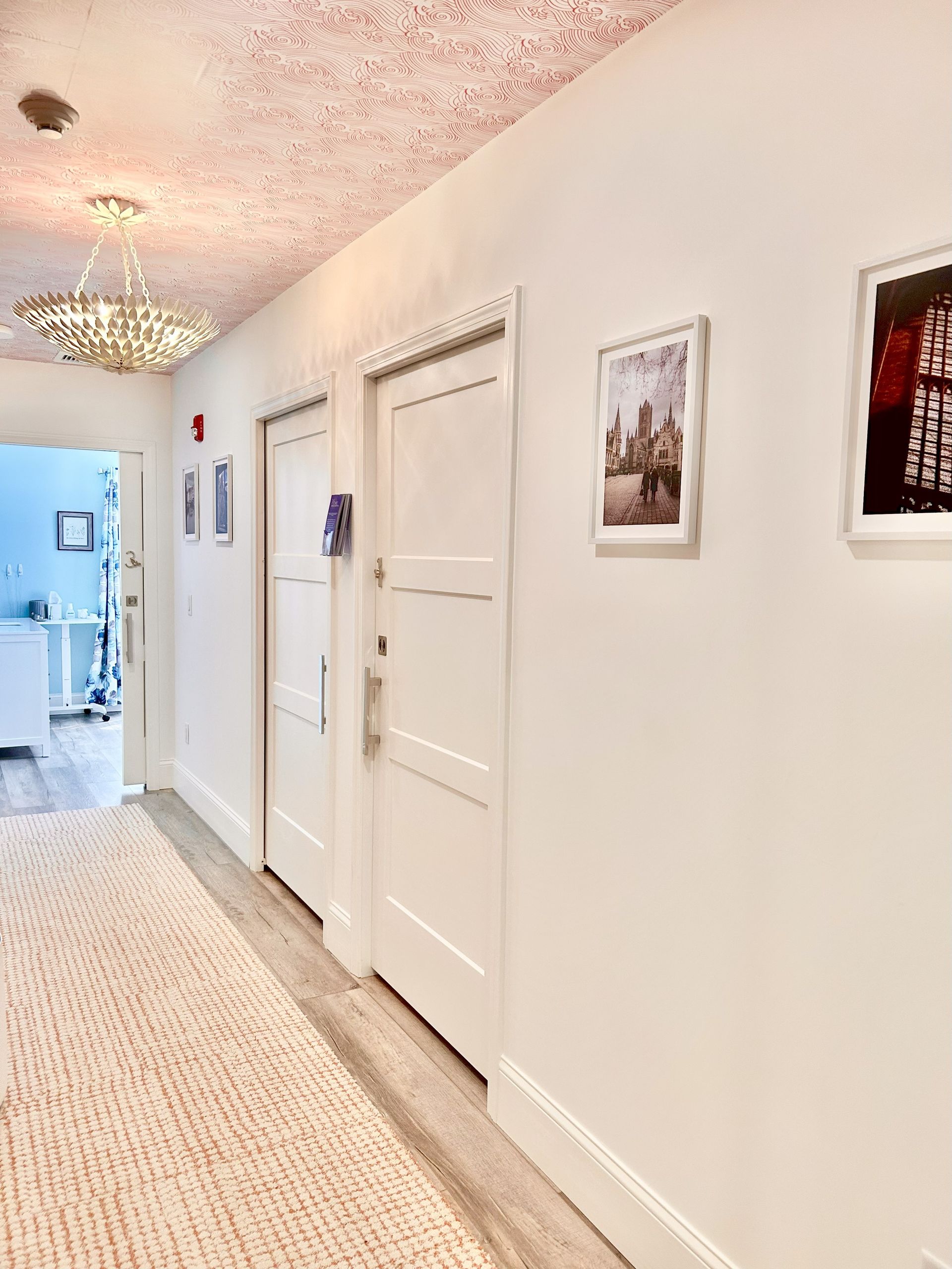 Hallway with white doors, artwork, and a textured rug. Bright lighting, white walls, and a view down a corridor.