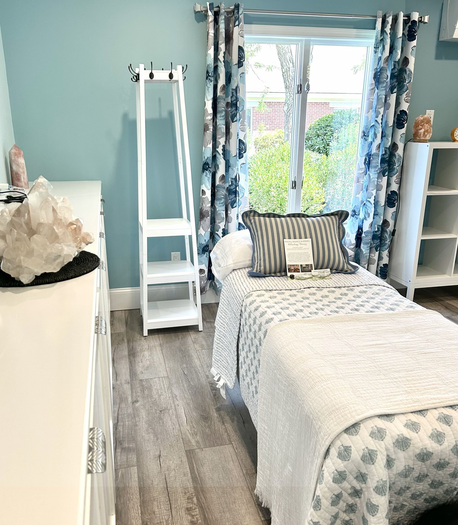 A serene room with a massage table, blue walls, and a window with floral curtains.